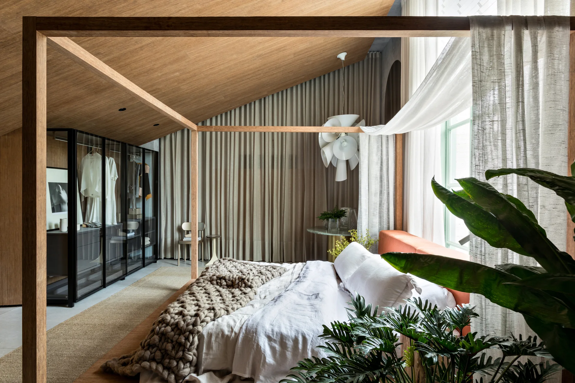 A modern bedroom with a wooden four-poster bed, sloped wood-paneled ceiling, full-length beige curtains, and a glass-fronted wardrobe.