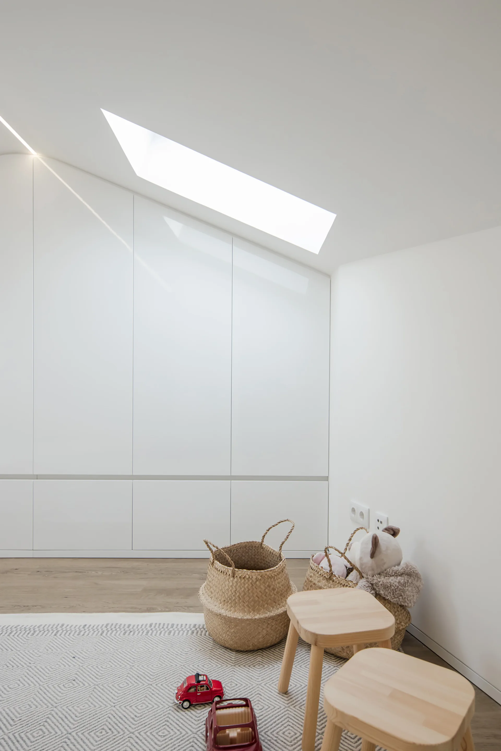 An attic playroom features a sloped ceiling with a skylight, white built-in cabinetry, two wooden stools, and woven toy baskets on a patterned rug.