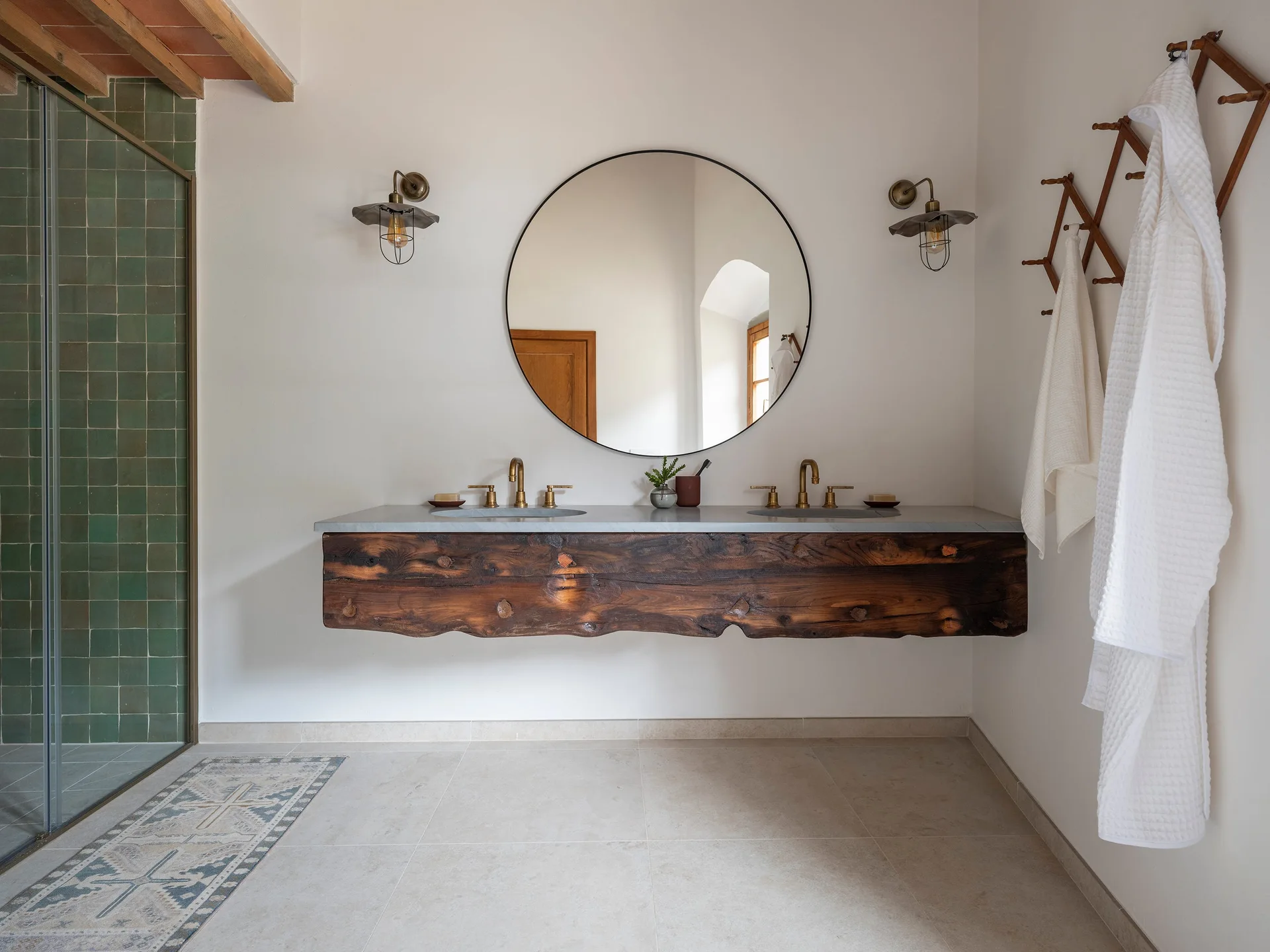 A rustic bathroom features a floating live-edge wood vanity with brass faucets under a large round mirror and metal wall sconces.