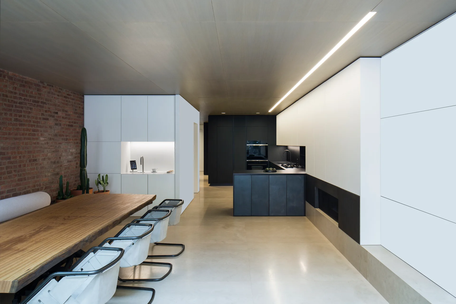 A minimalist open-plan kitchen features a wood paneled ceiling with a linear light, a live-edge dining table, white cabinetry, and an exposed brick wall.