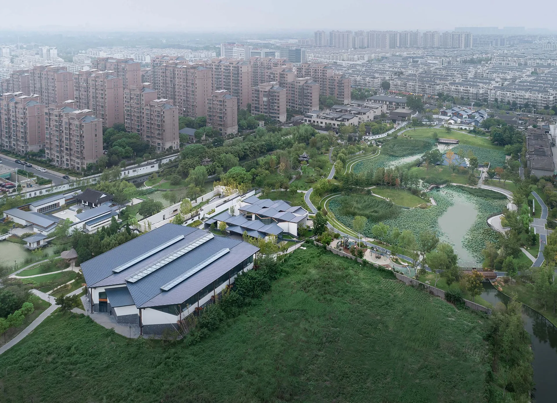 An aerial view of a modern building with dark pitched roofs situated next to winding paths and ponds in an urban park.
