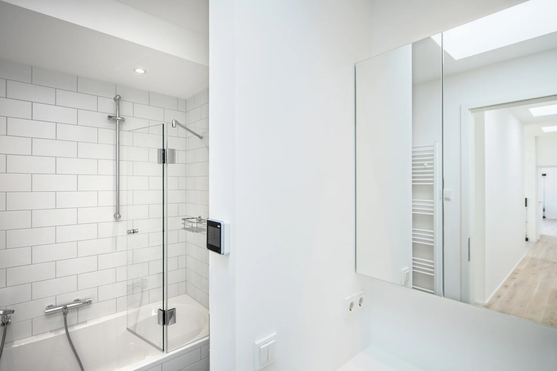A minimalist white bathroom featuring a bathtub with subway tile, a glass shower partition, and a large wall mirror.
