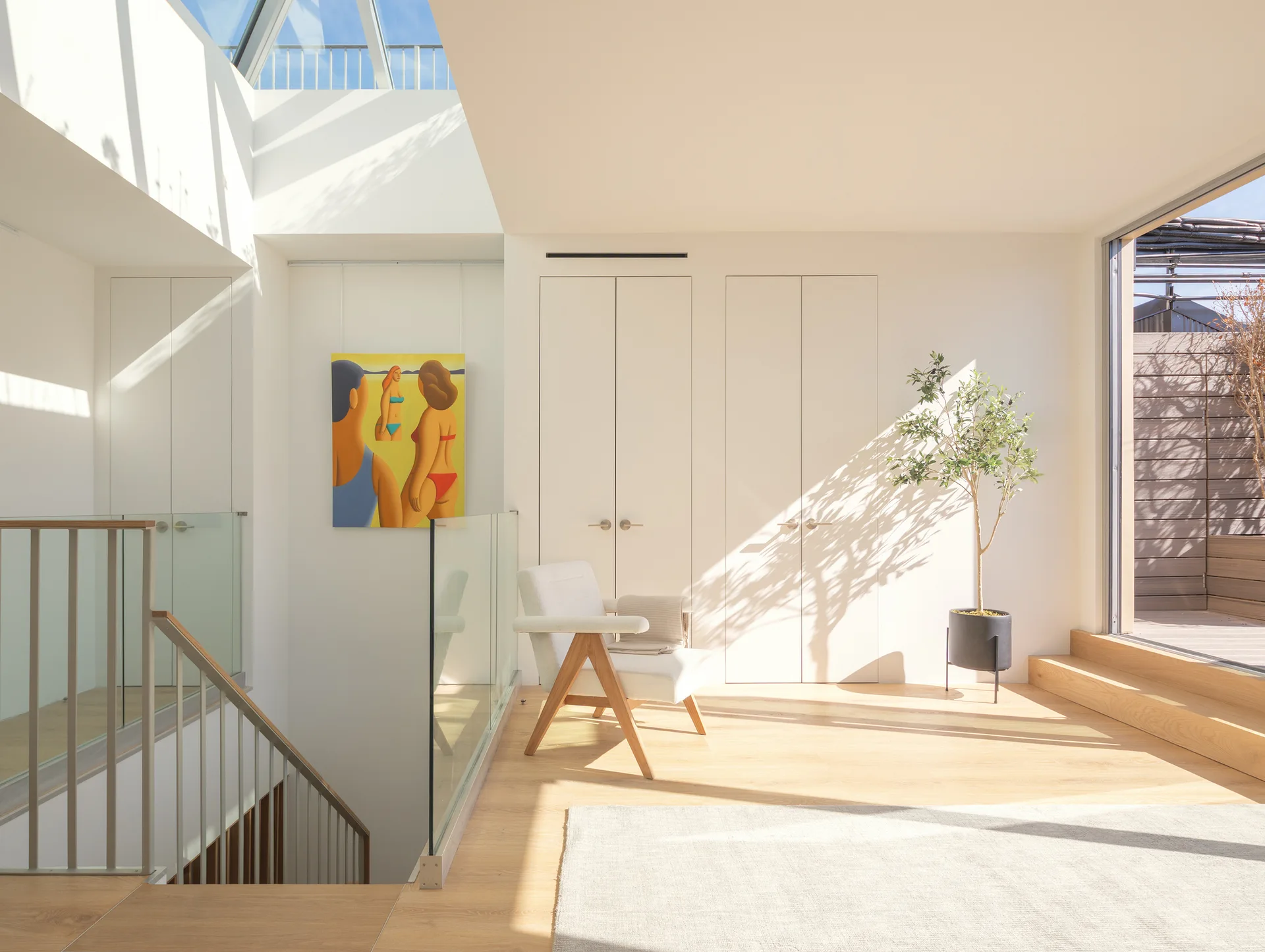 A bright staircase landing features a white armchair, a potted tree, and a glass railing illuminated by a large overhead skylight.