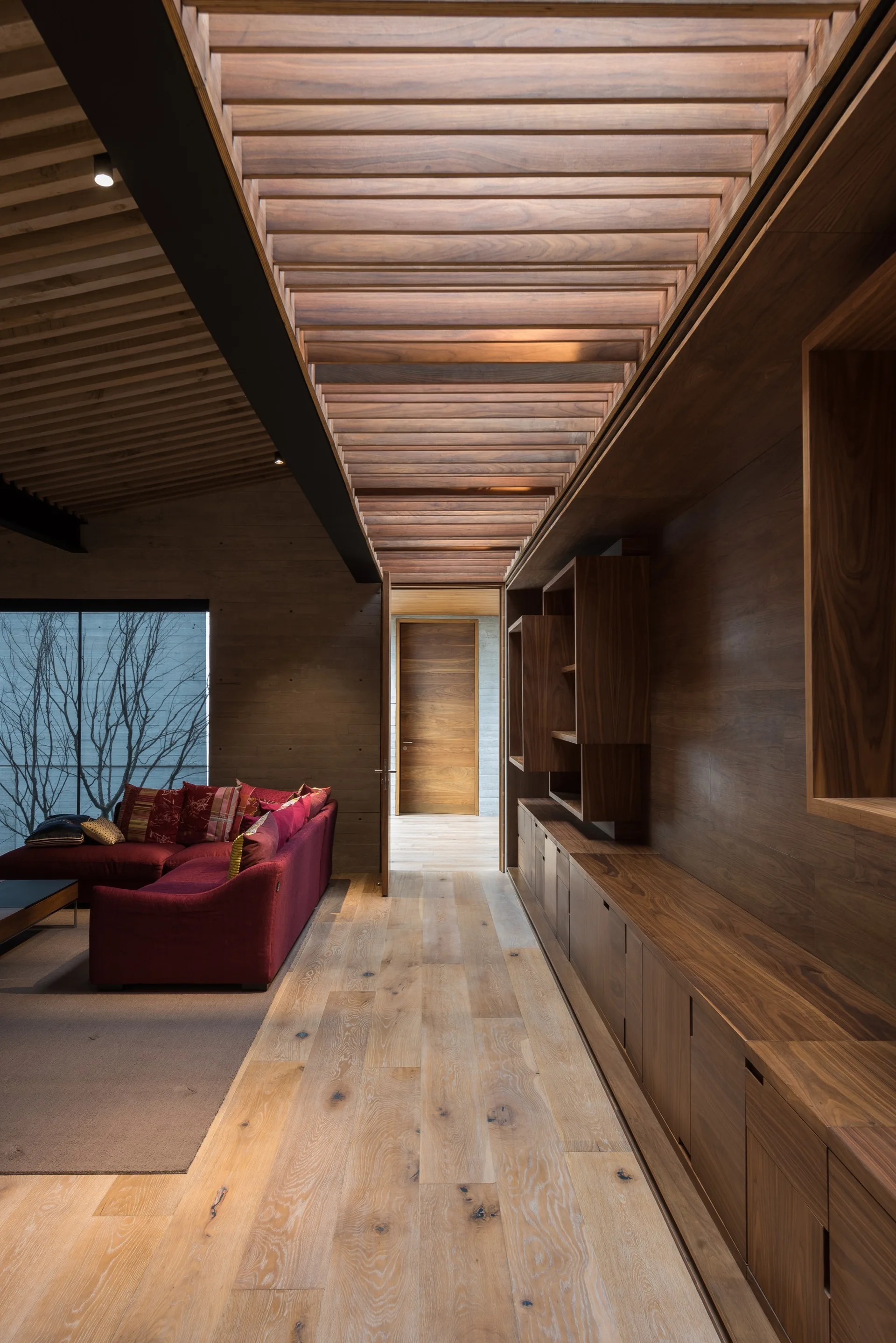 A modern living area featuring a deep red sectional sofa, wood paneled walls, and a distinctive slatted timber ceiling skylight.