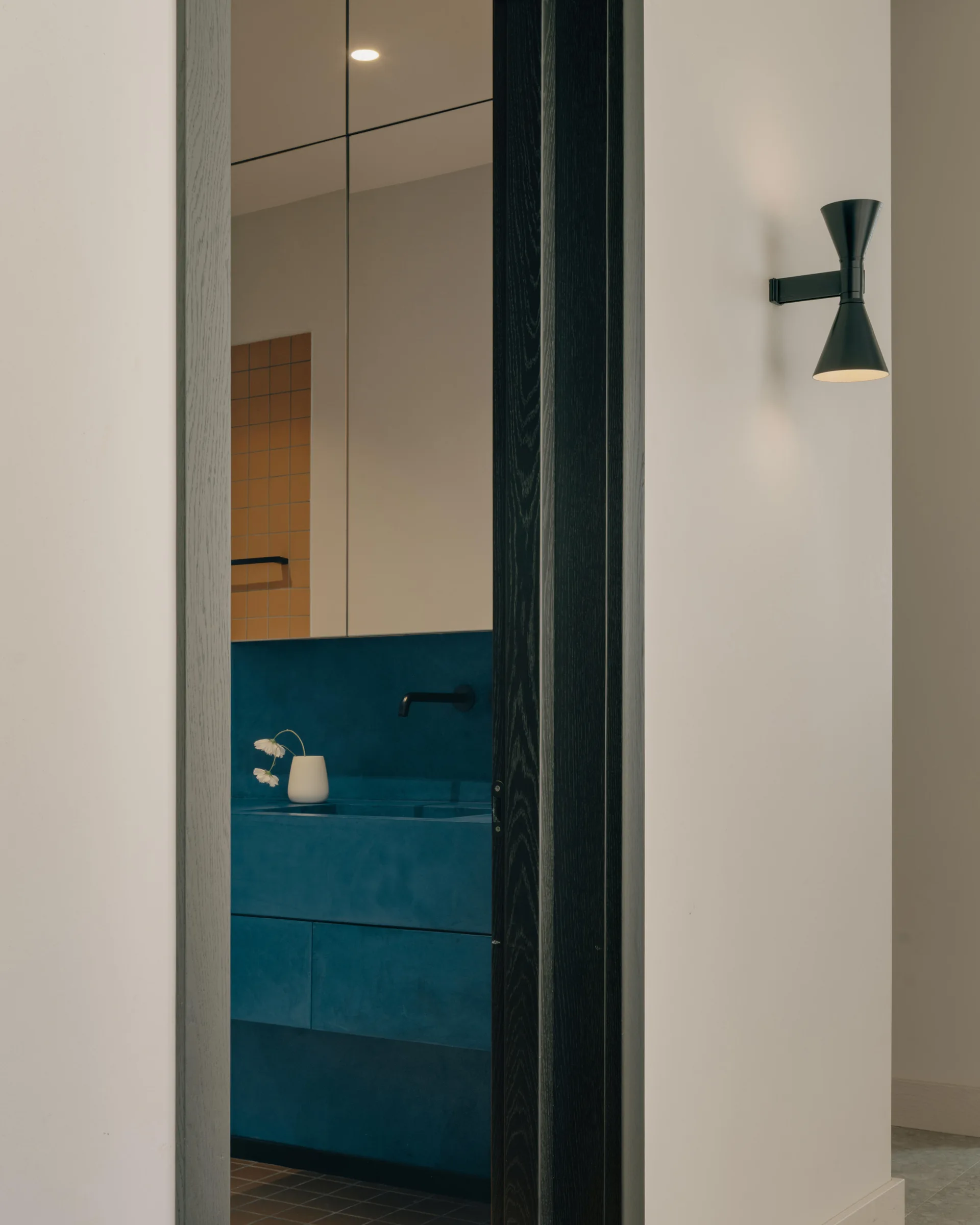A view through a doorway reveals a bathroom with a matte blue vanity and yellow accent tiles, with a black sconce mounted on the outside wall.