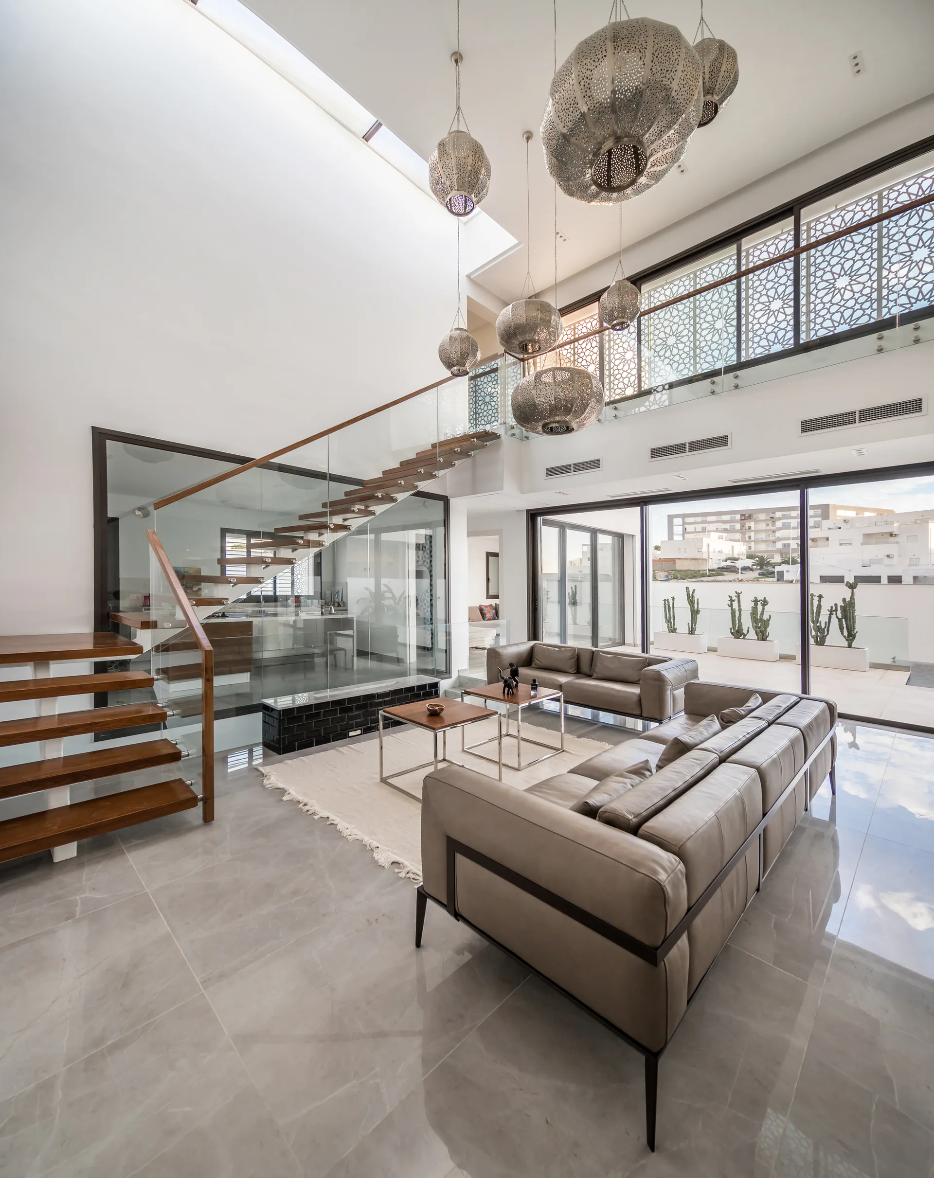 A modern double-height living room featuring grey leather sofas, a floating wooden staircase with glass railings, and hanging globe pendant lights.
