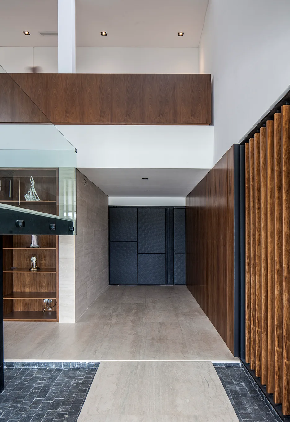 A modern entryway with high ceilings, featuring vertical wood slat paneling on the wall, large stone floor tiles, and built-in wooden shelving.