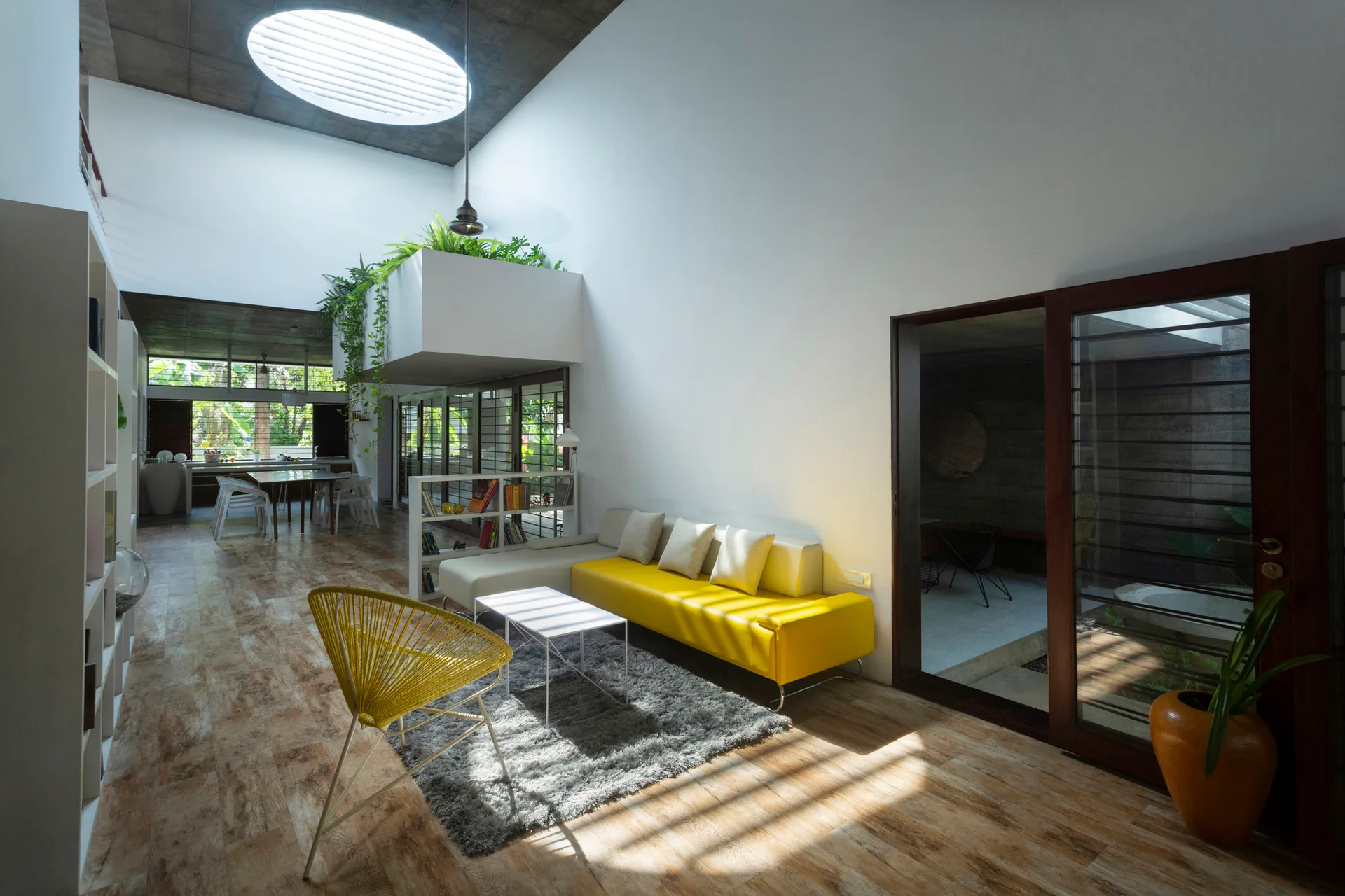 A modern open-plan living room featuring a yellow sectional sofa, a shaggy grey rug, and a large circular skylight in a concrete ceiling.