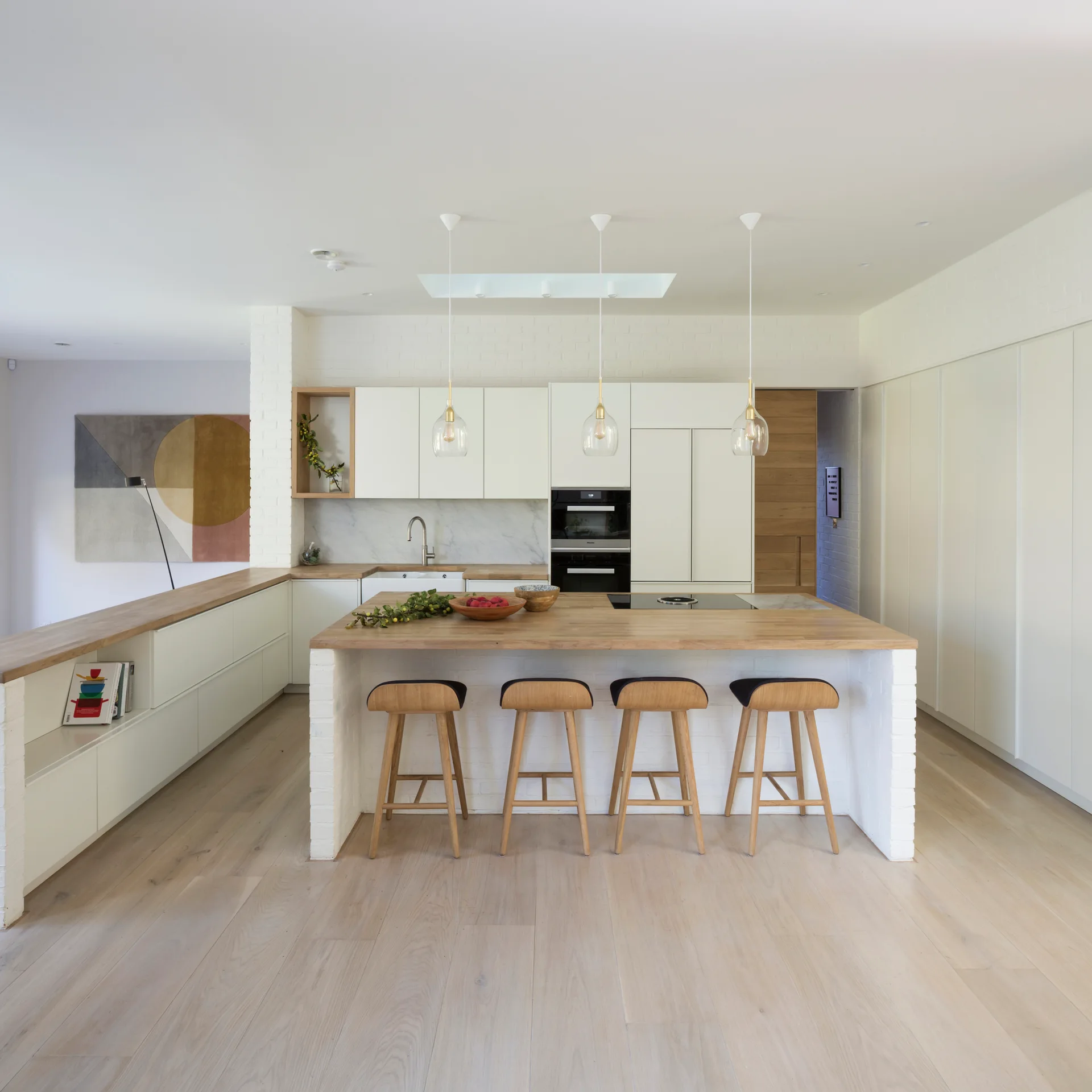 A modern Scandinavian kitchen features white cabinetry, a light wood-topped island with four matching wooden bar stools, and glass pendant lights.