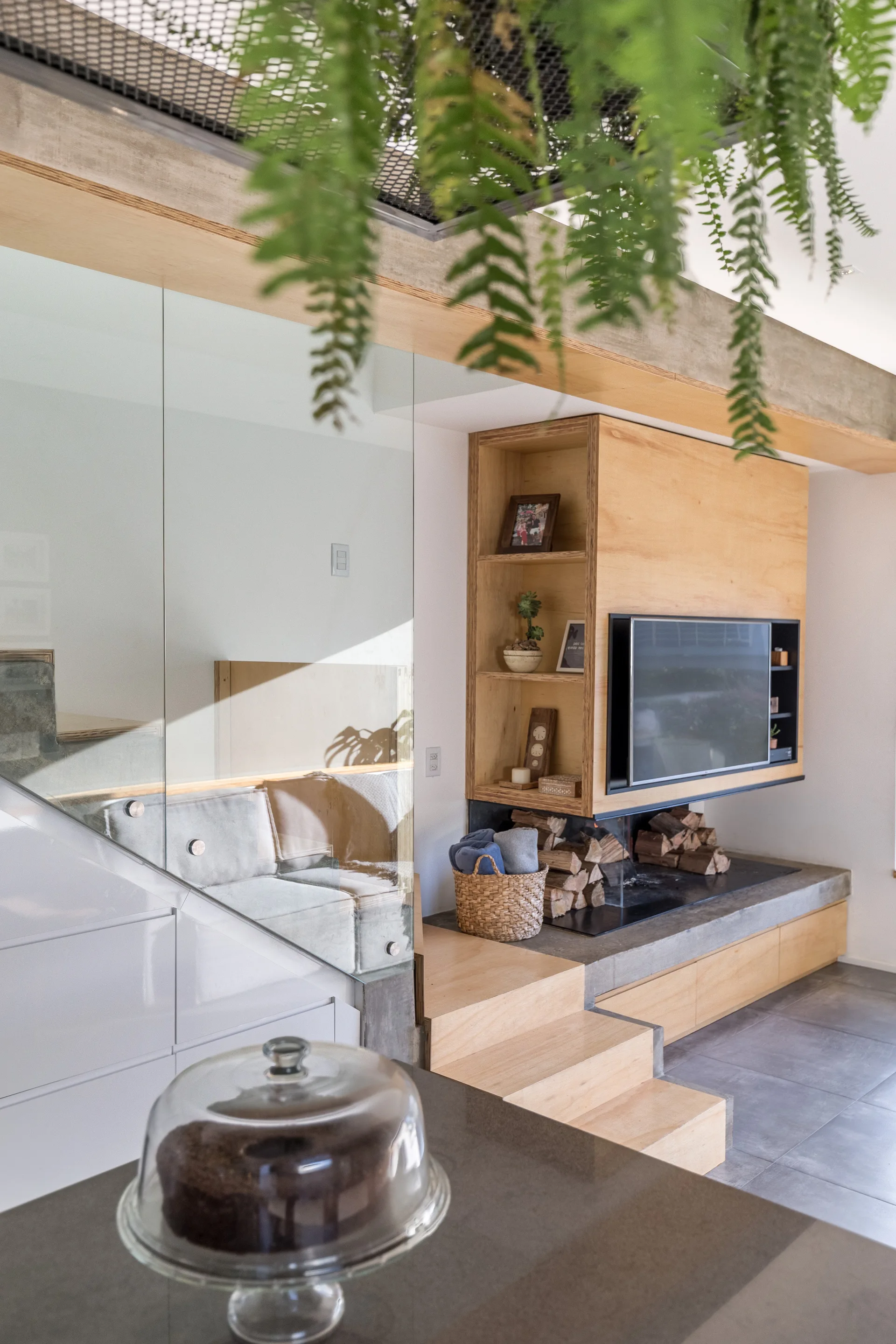 A modern living area featuring a custom light wood media cabinet, an integrated low concrete fireplace hearth, and a transparent glass stair wall.