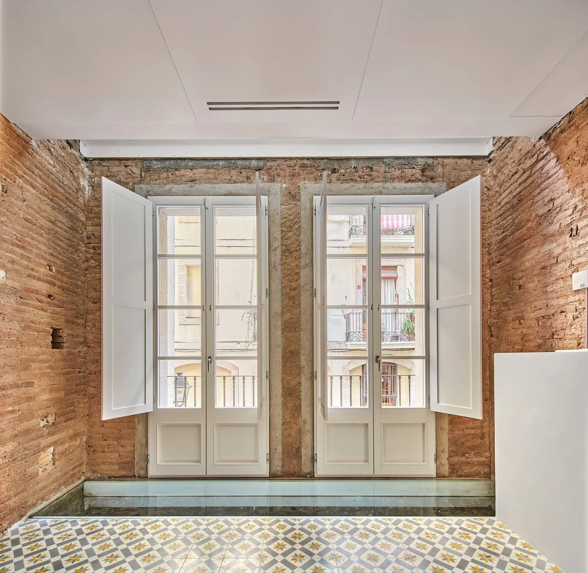 An interior space featuring raw exposed brick walls, two large white French windows with open shutters, and geometric patterned floor tile.