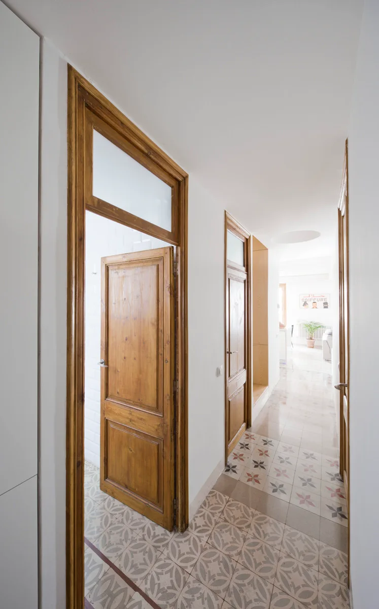 A long white hallway features natural wood doors with glass transom windows and a floor covered in geometric patterned tiles.