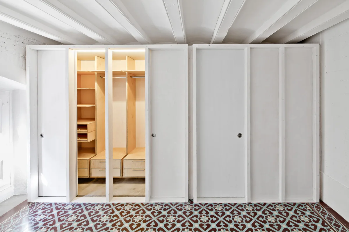 A custom white closet system with sliding doors revealing light wood shelves and drawers sits on patterned geometric floor tiles below a beamed ceiling.