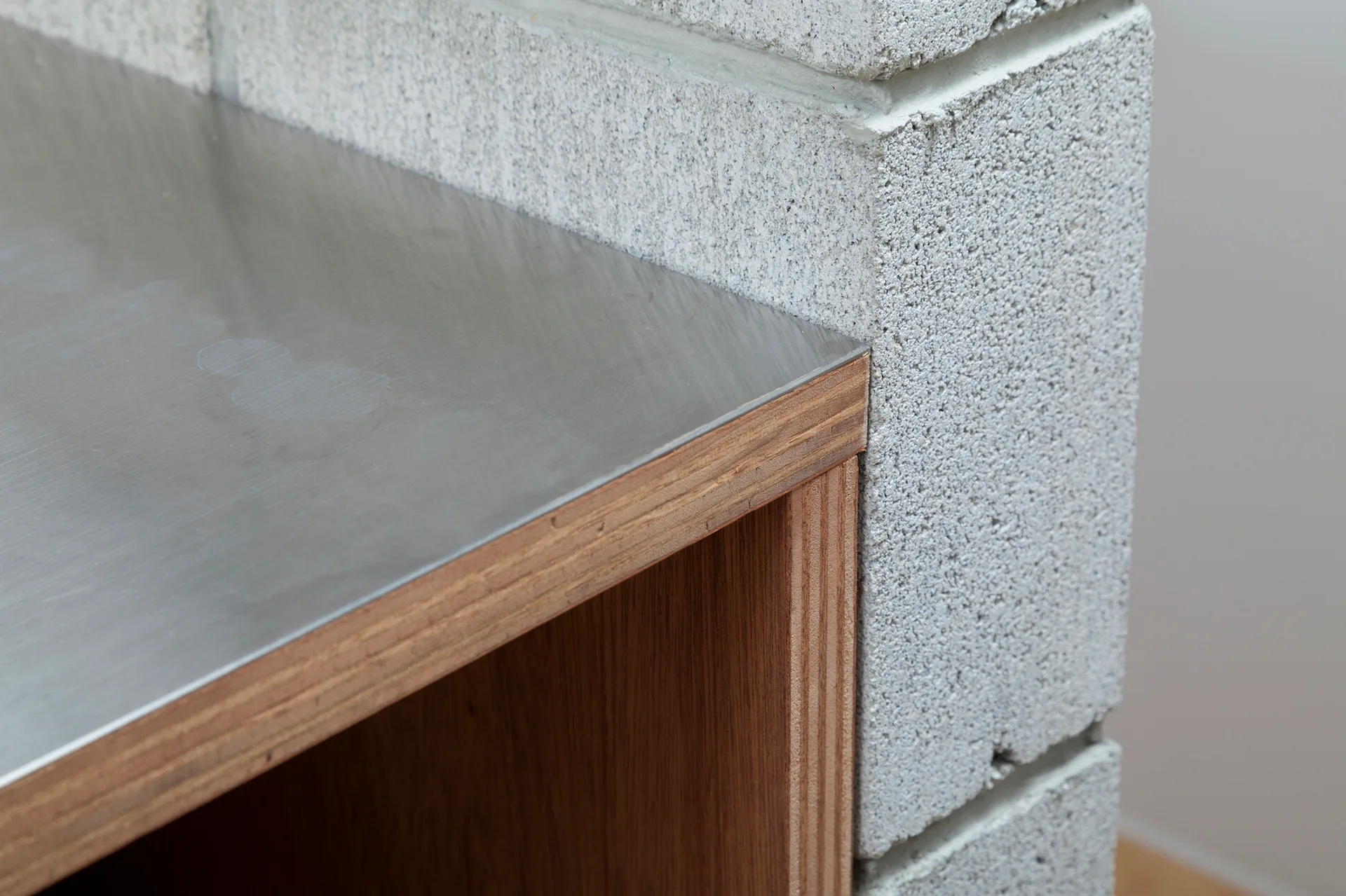 A close-up of a stainless steel countertop with an exposed plywood edge abutting a grey concrete block wall.