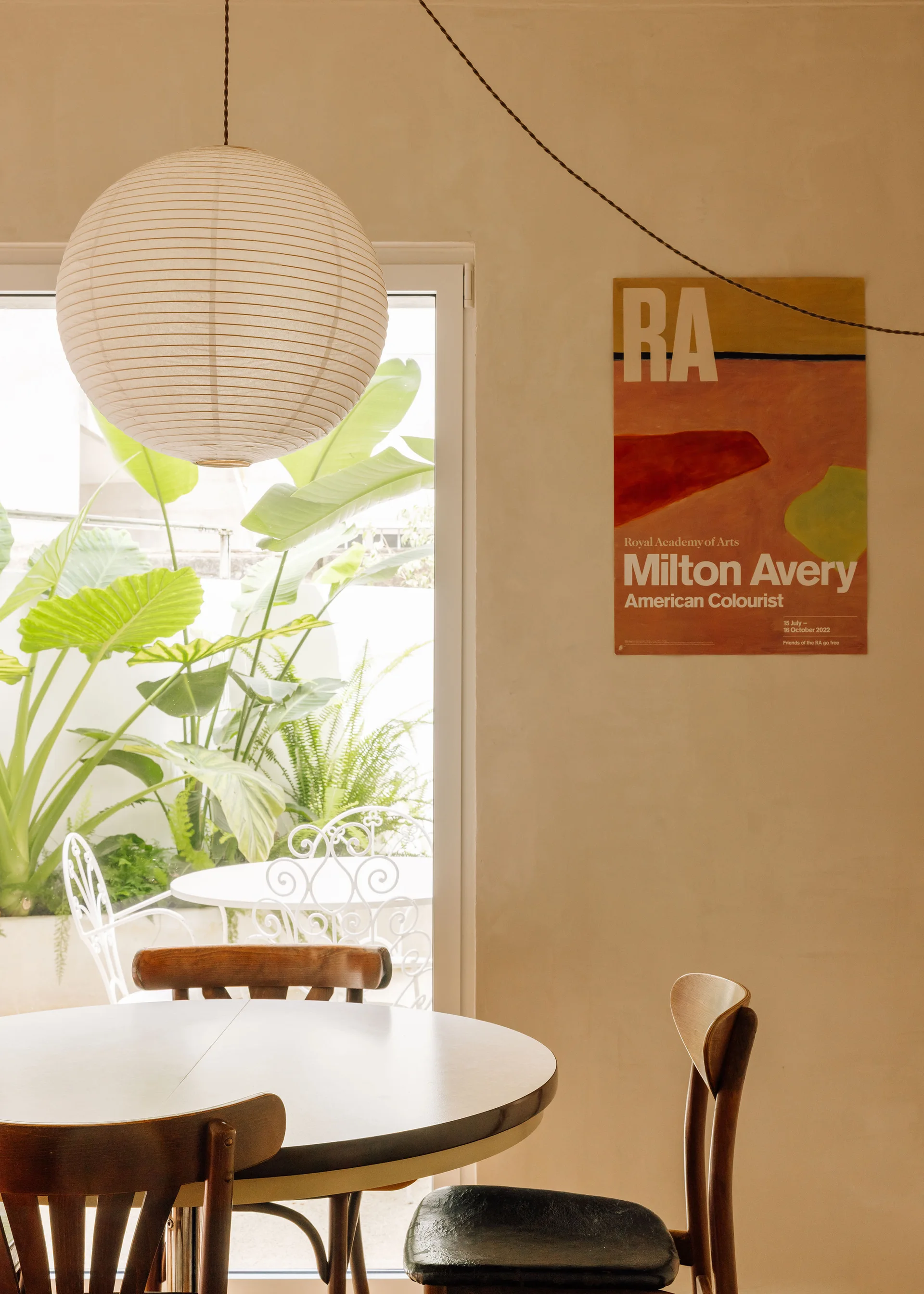 A bright dining room featuring a round wooden table, bentwood chairs, a large paper globe pendant light, and a colorful art print on a plaster wall.