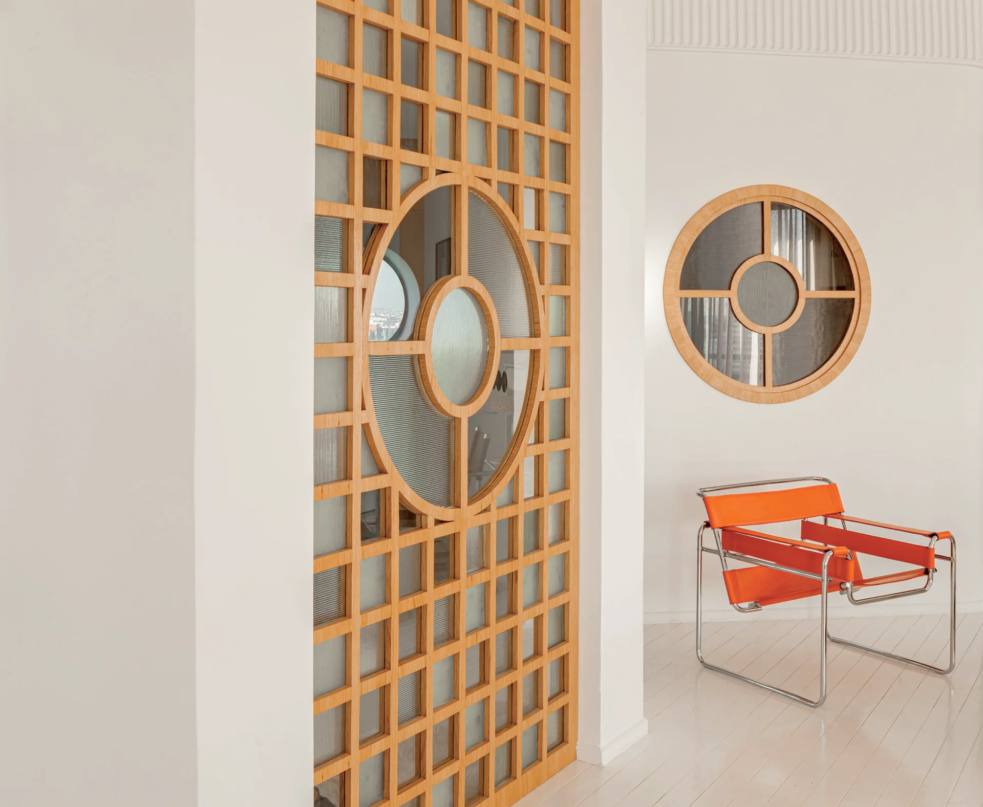 A light wood grid room divider with fluted glass stands near a circular wall window and a bright orange metal frame lounge chair.