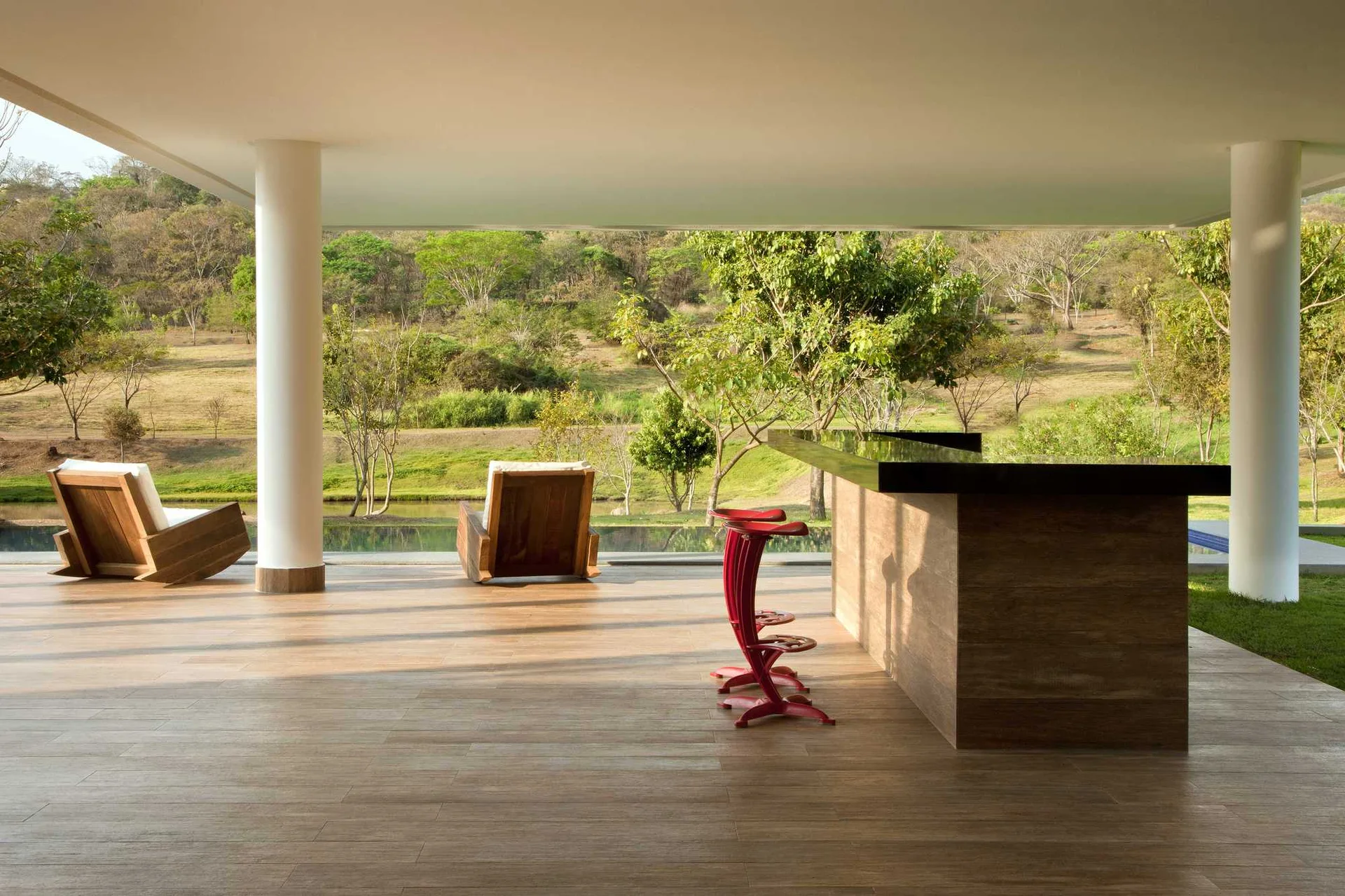 A covered modern patio with wood flooring, a wood bar counter with red stools, and lounge chairs overlooking a grassy landscape.
