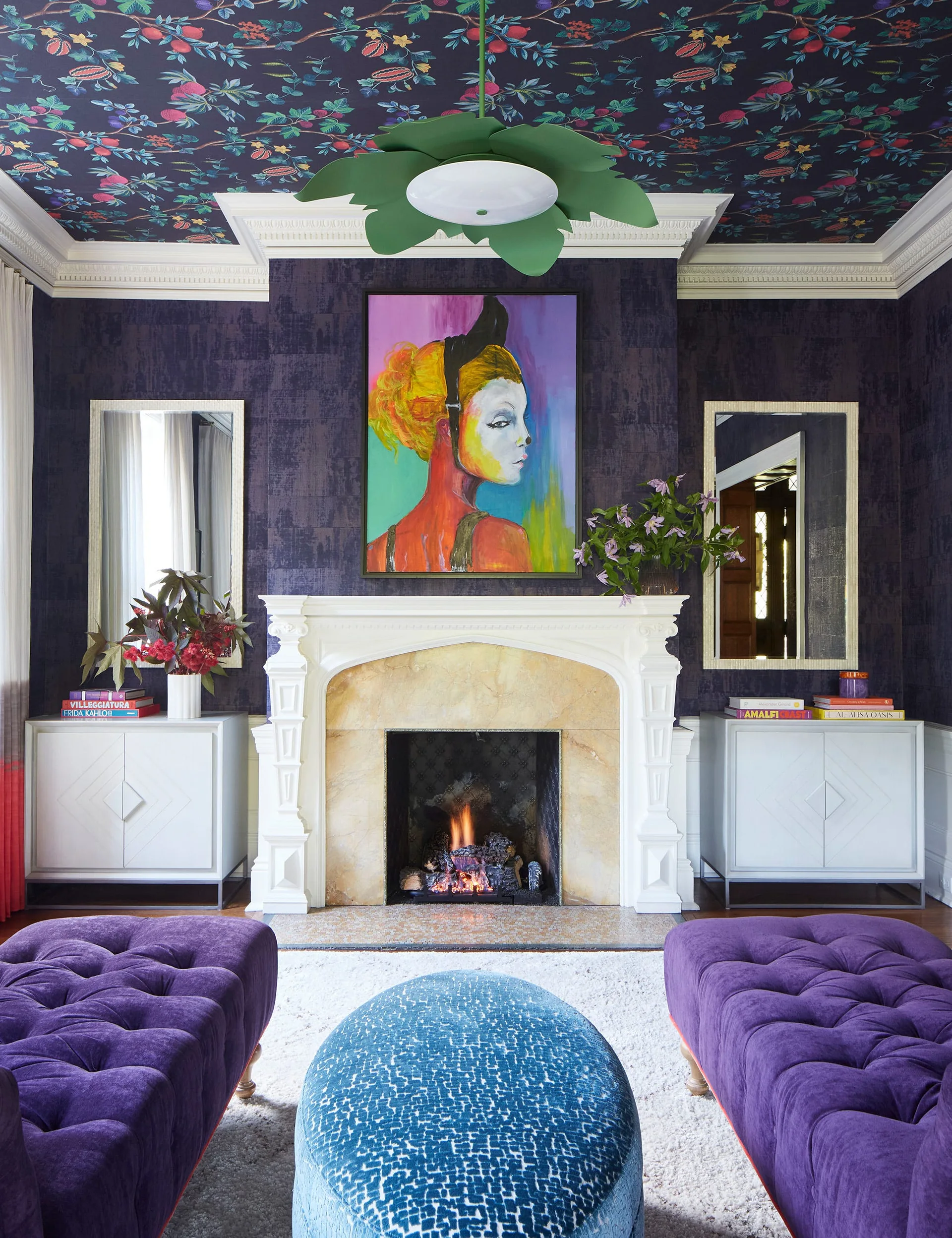 A living room with a white fireplace mantel against purple textured walls beneath a botanical patterned ceiling, with purple tufted ottomans.