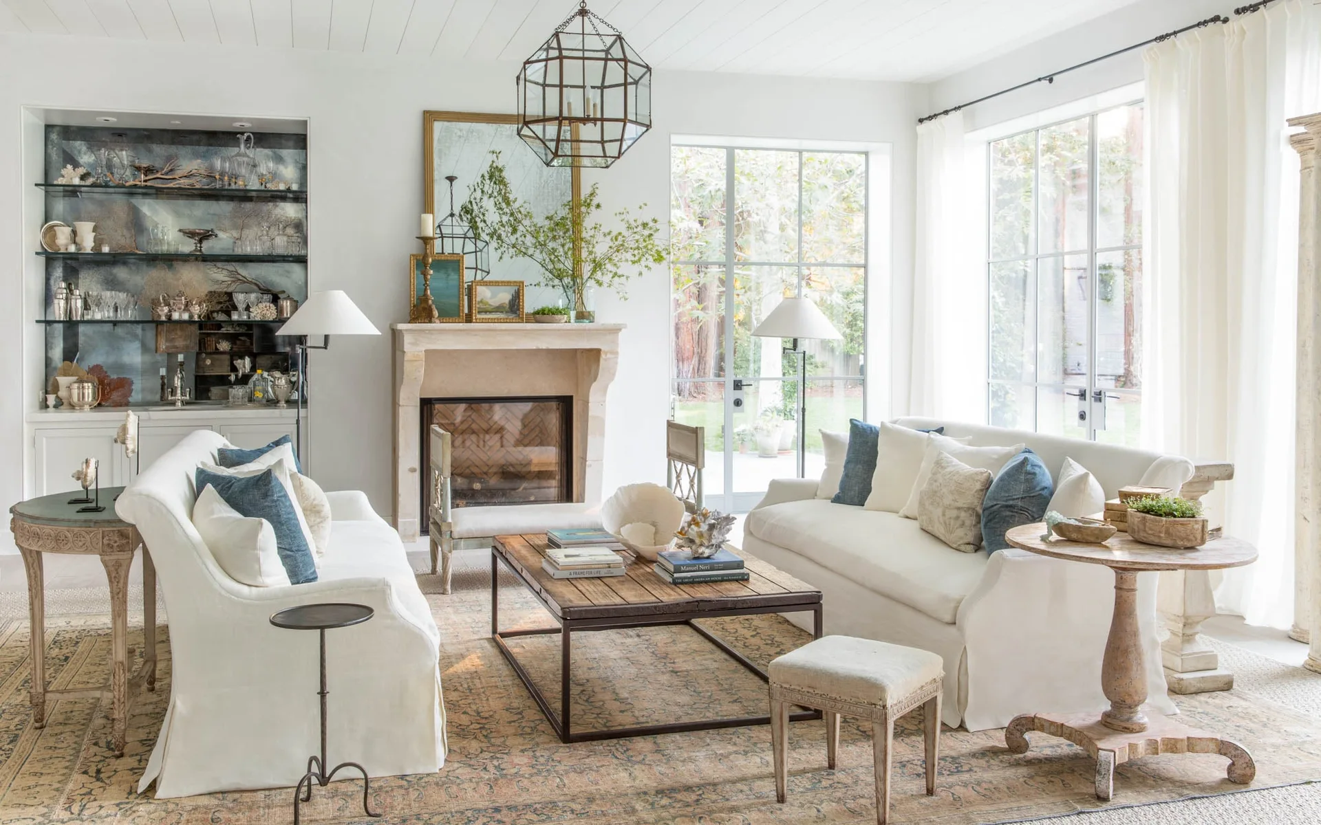 A bright living room features two white slipcovered sofas facing a rustic wooden coffee table, a stone fireplace, and large grid windows.