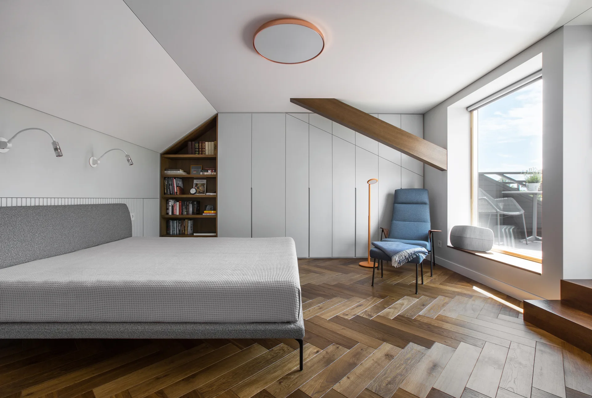 A contemporary attic bedroom featuring a grey bed, herringbone wood flooring, built-in wardrobes, and a blue armchair by a bright window.