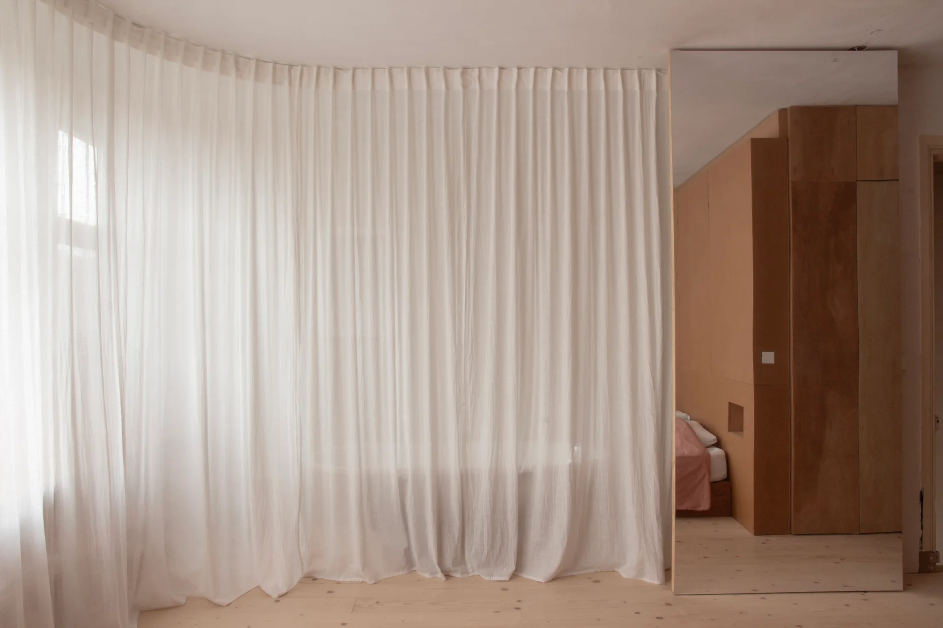 Floor-to-ceiling white sheer curtains hang in a minimalist bedroom next to a large mirror reflecting a plywood wardrobe and bed.