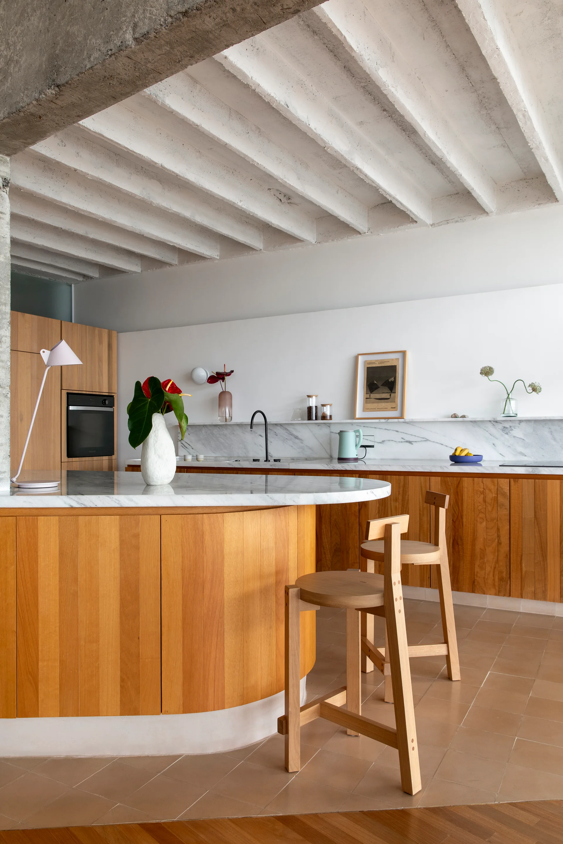 A minimalist kitchen features a curved wooden island with a marble countertop, matching wood stools, terracotta floor tiles, and white exposed ceiling joists.