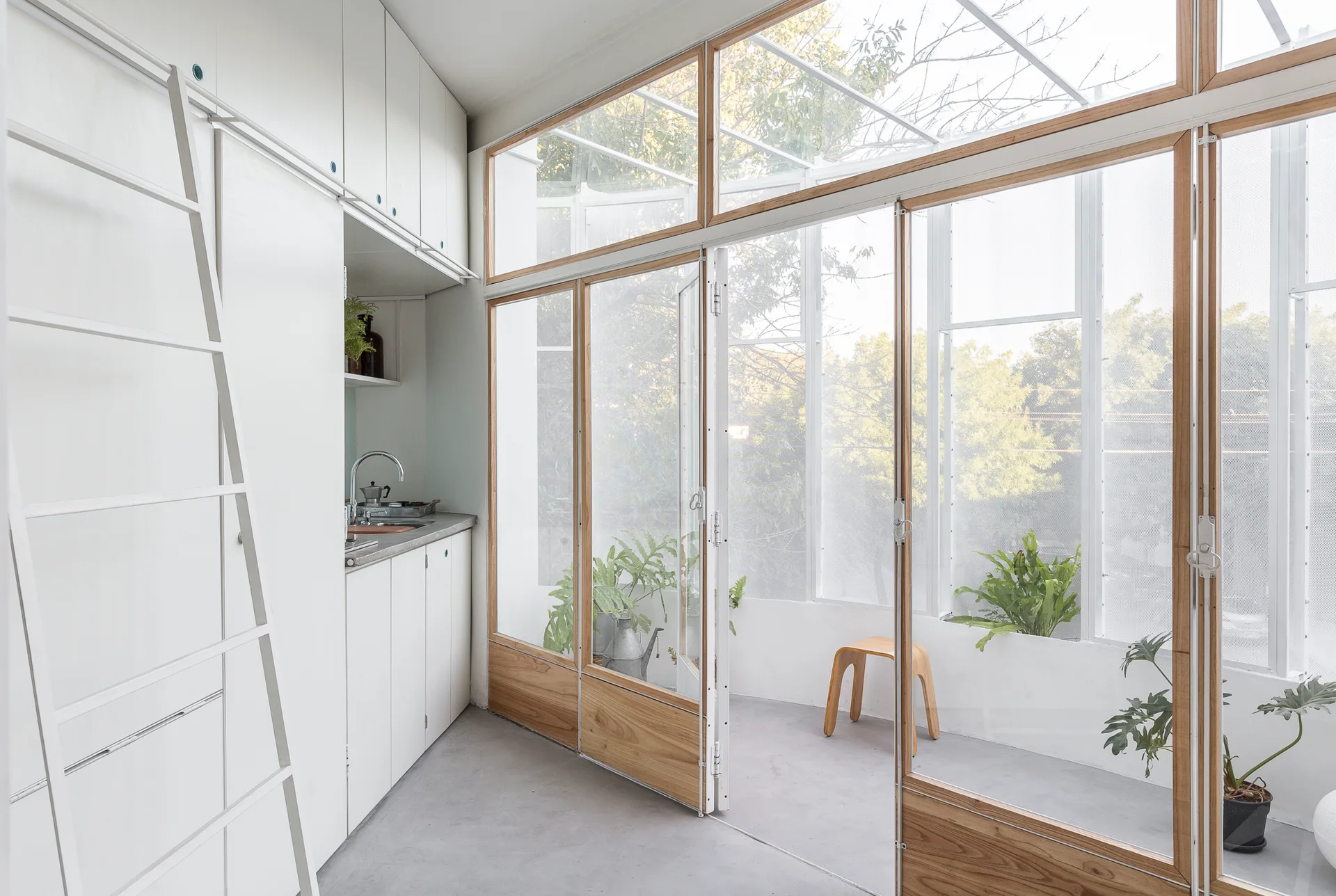 White built-in kitchen cabinets and a leaning ladder sit next to wood-framed glass doors leading to a bright sunroom with concrete flooring.
