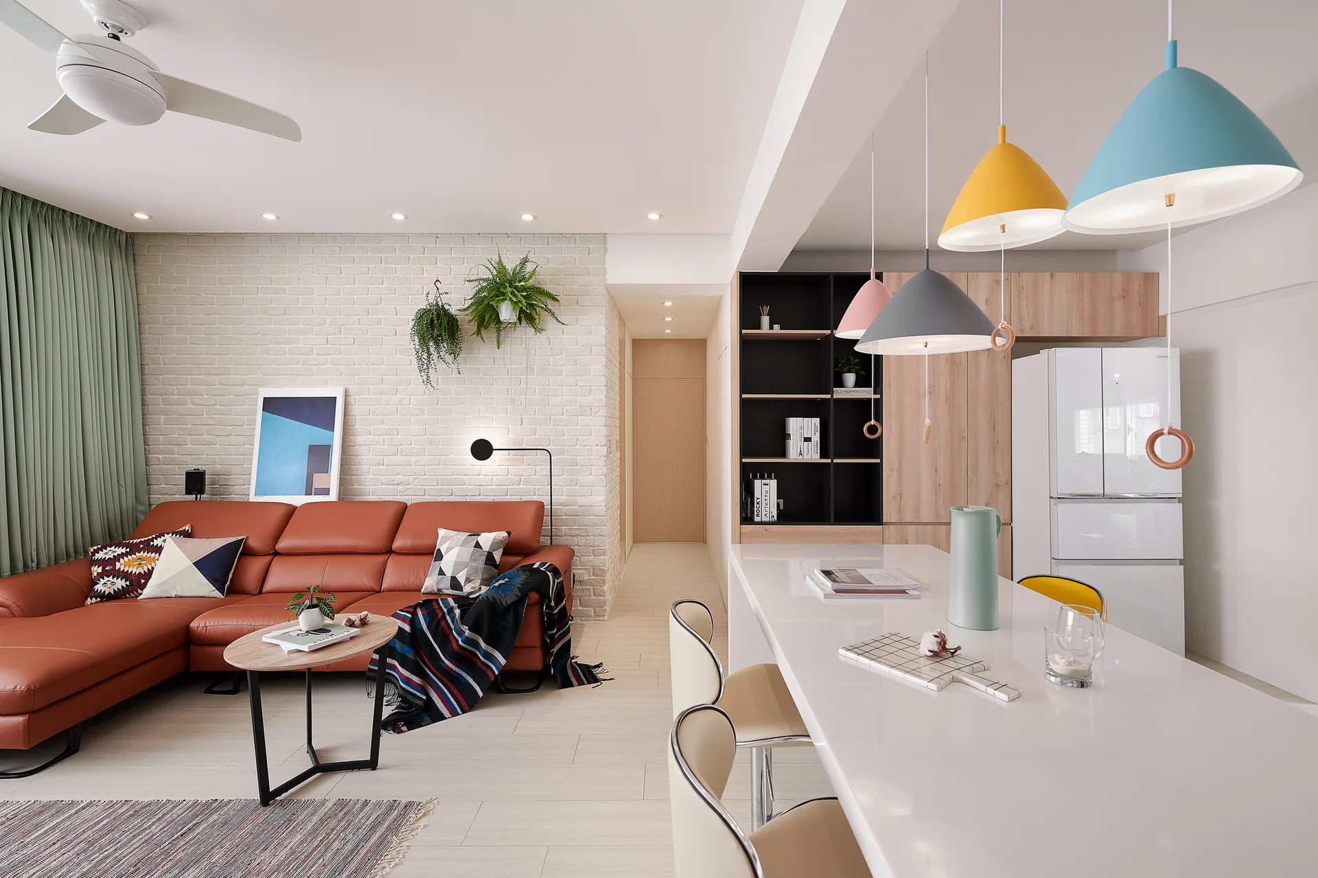 An open living space features a rust-colored leather sofa against a white brick wall and a white kitchen island with multi-colored pendant lights.