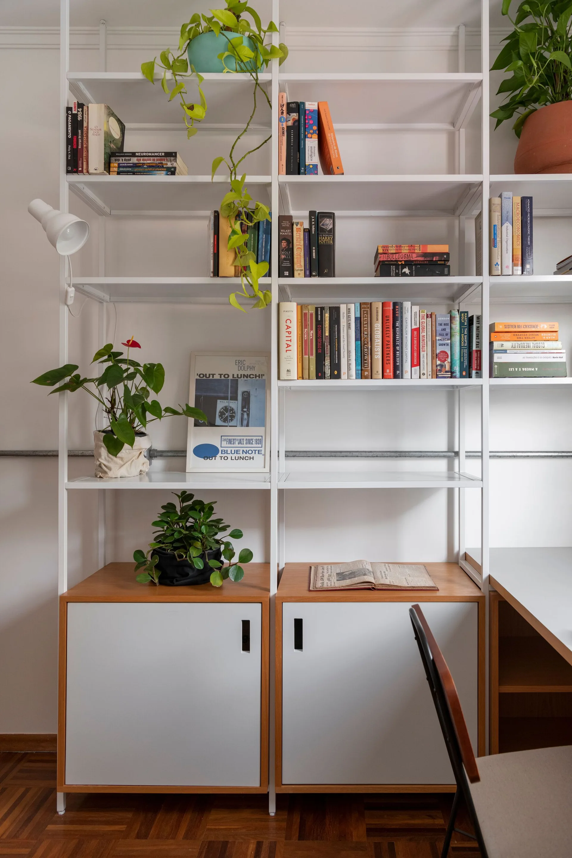 A white metal modular shelving unit filled with books and potted plants sits above light wood base cabinets on a parquet floor.