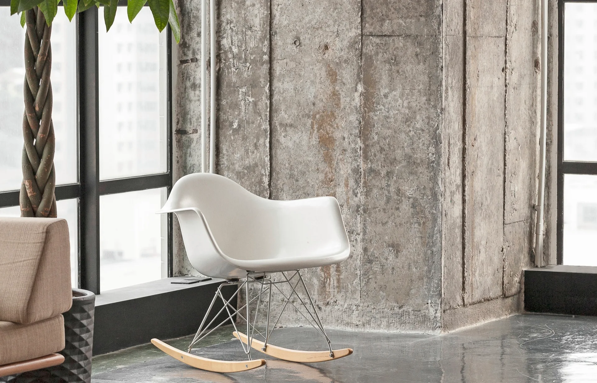 A white mid-century modern rocking chair stands near a large window in a room with rough exposed concrete walls.