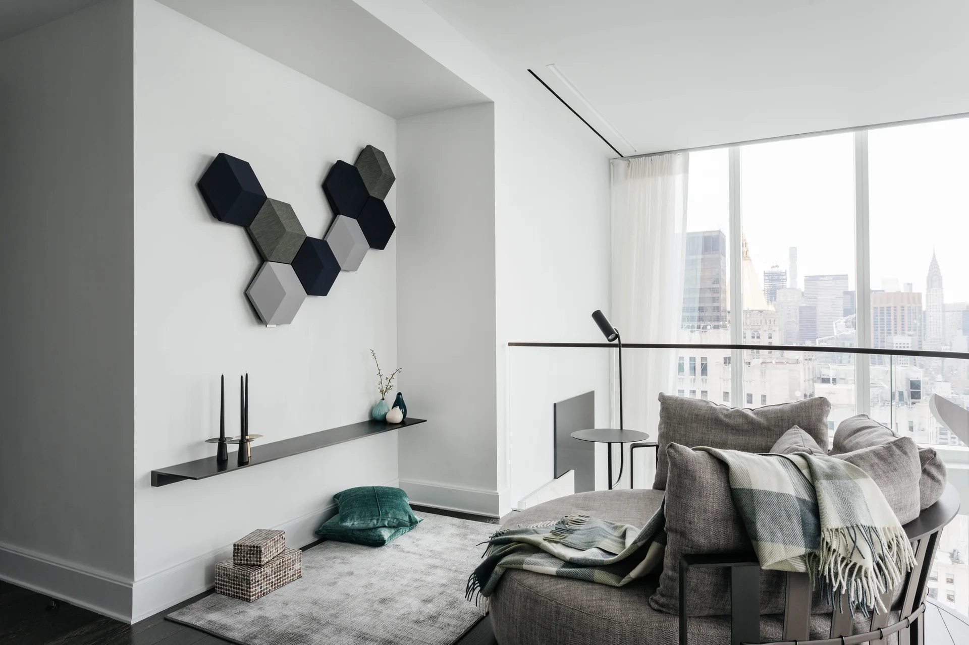 A contemporary living room features a circular grey sofa, a long black floating shelf, and geometric hexagonal wall panels against white walls.