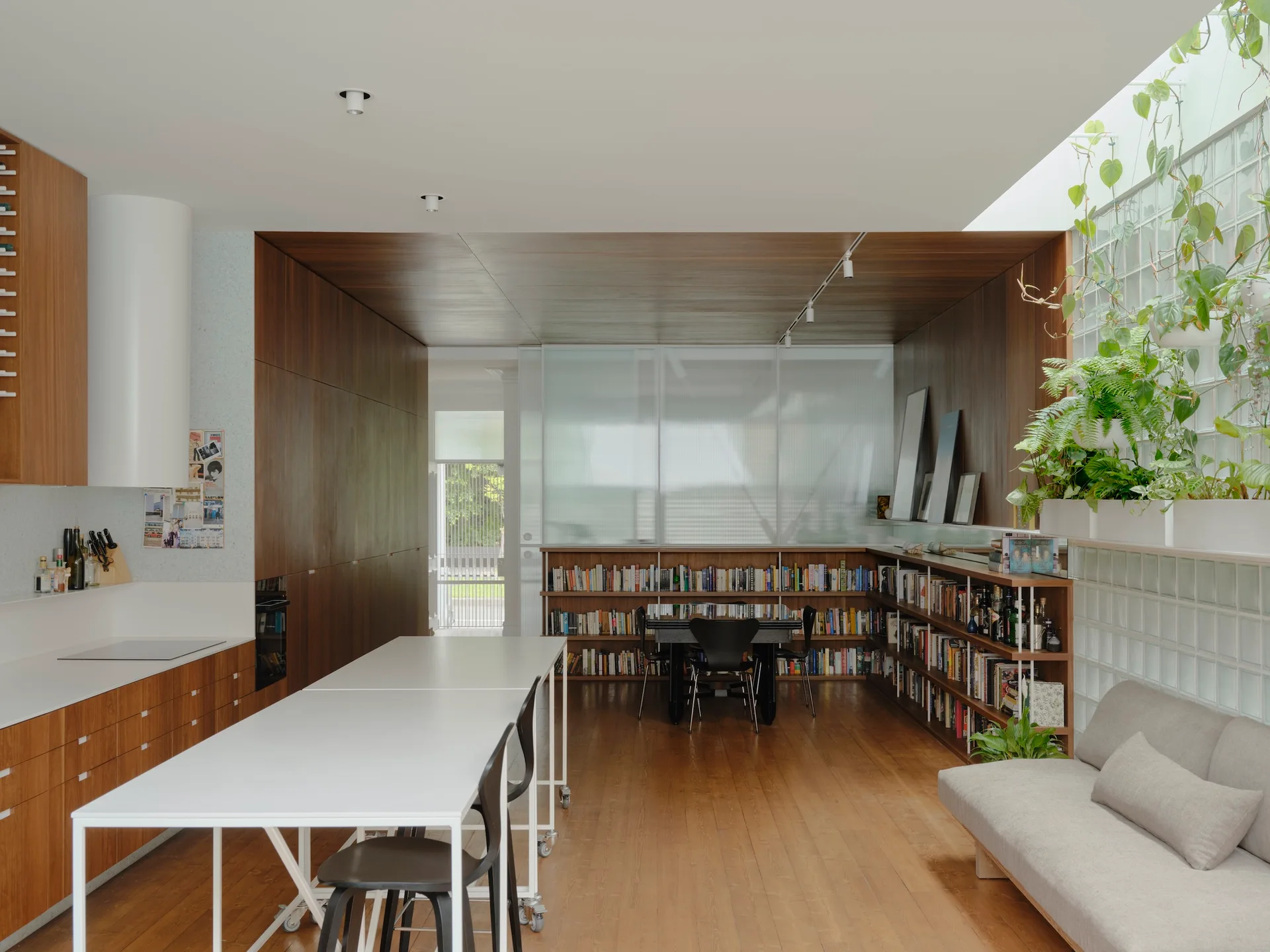 A spacious modern kitchen and living area featuring warm wood paneling, a long white table on wheels, a glass block wall, and low bookshelves.