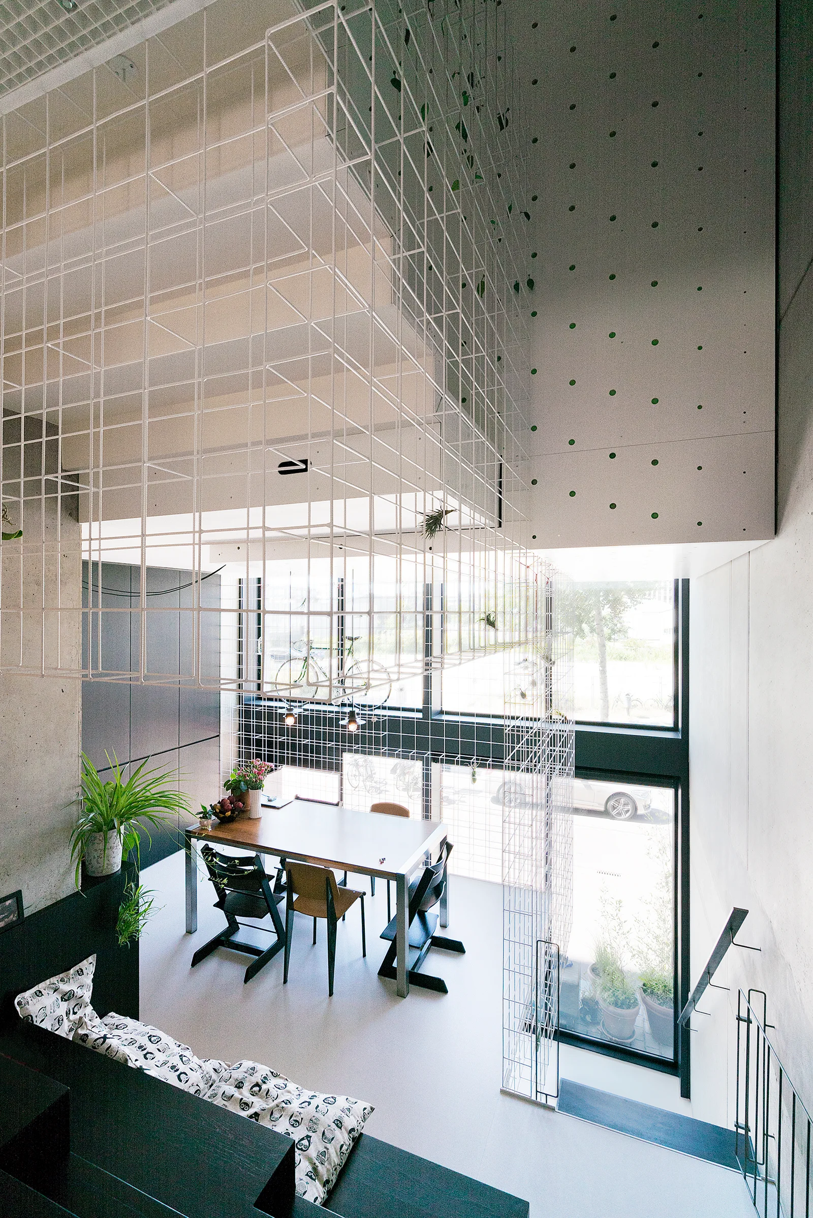 A modern double-height dining room features a large rectangular table, a suspended white wire grid structure, and tall windows showing an outdoor street view.