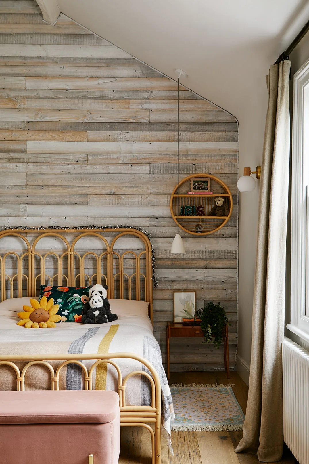 A children's bedroom features a rattan bed frame, a horizontal wood-paneled wall, a round rattan shelf, and a pink storage bench.