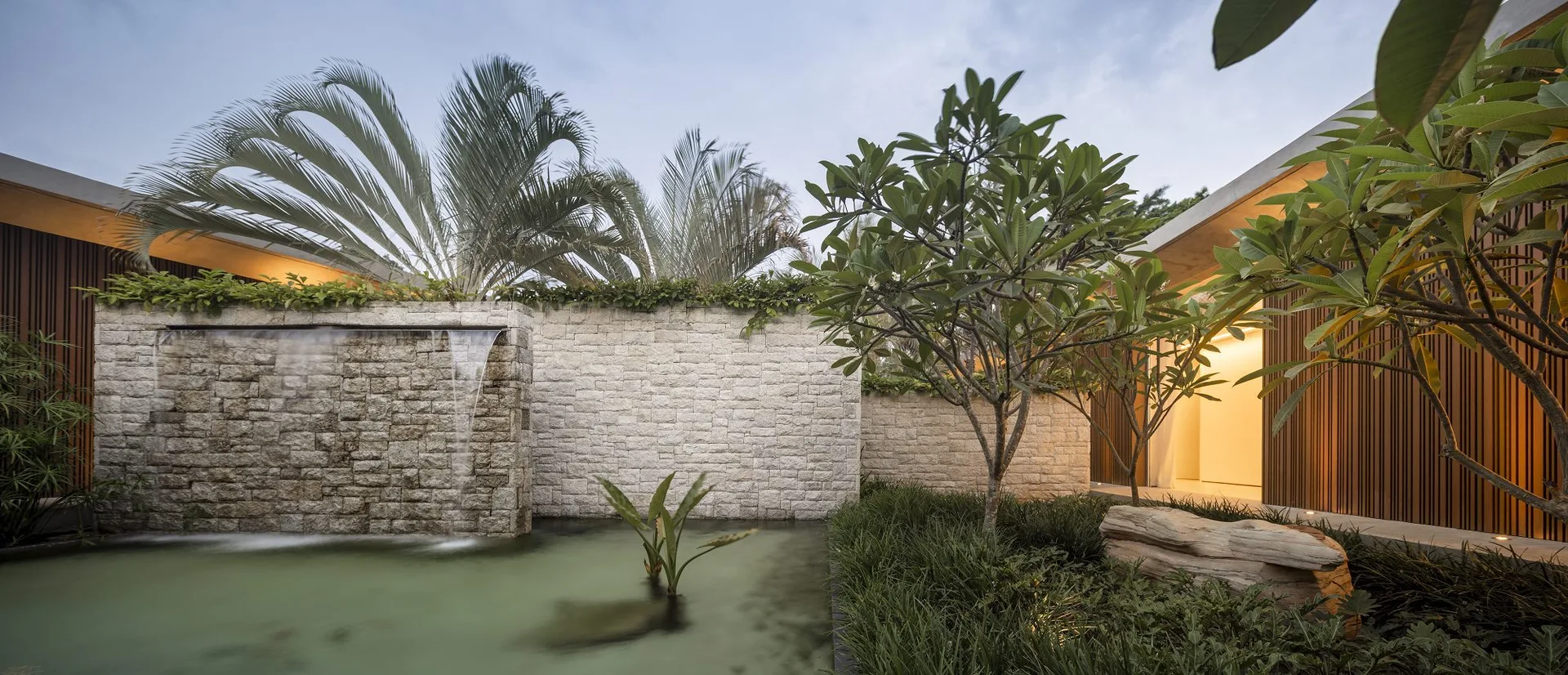 A modern outdoor garden featuring a stacked stone water wall cascading into a reflecting pool, surrounded by lush tropical plants and vertical wood cladding.