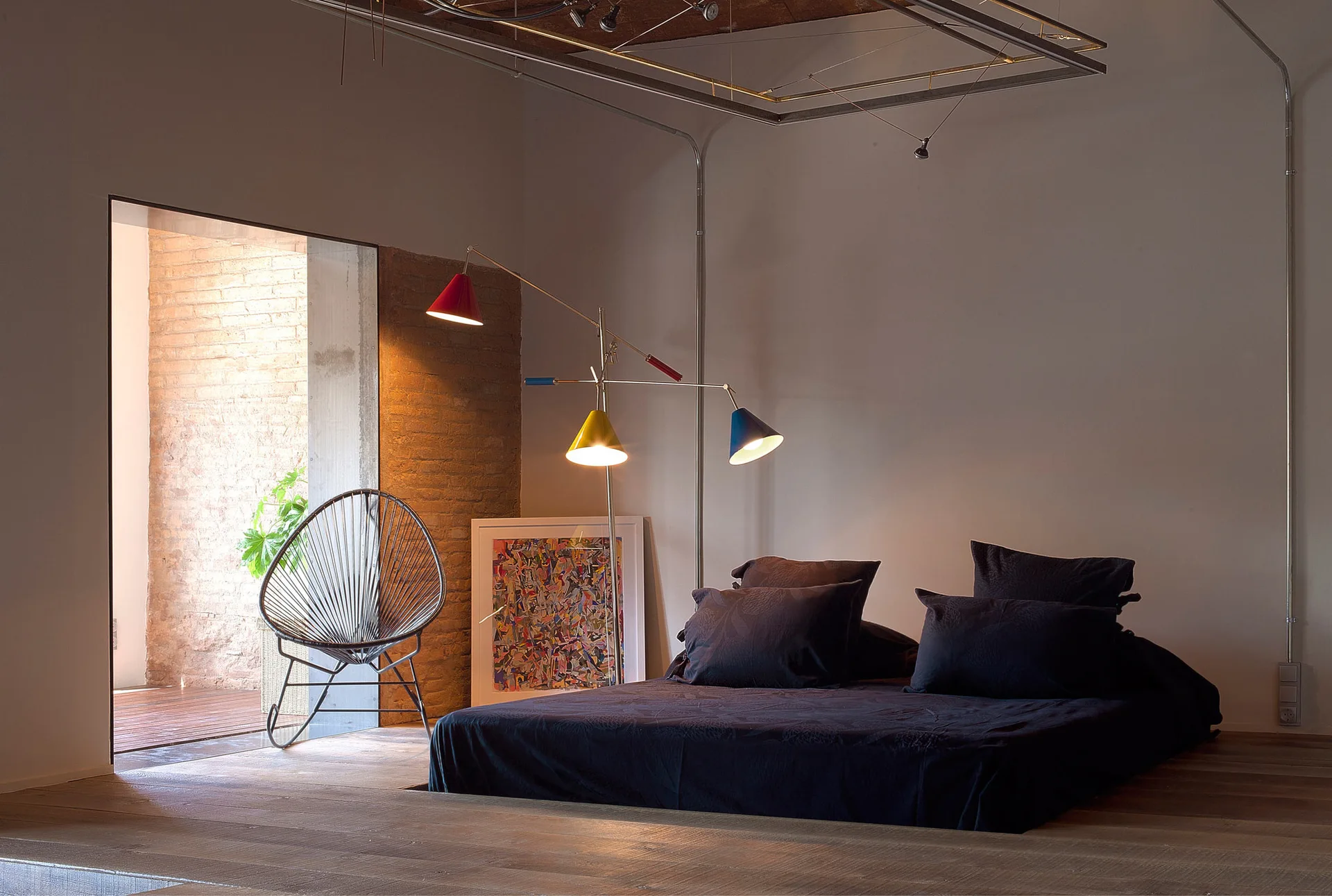 A low-profile dark bed on a wooden floor next to a colorful three-armed floor lamp and a metal wire chair near an exposed brick doorway.