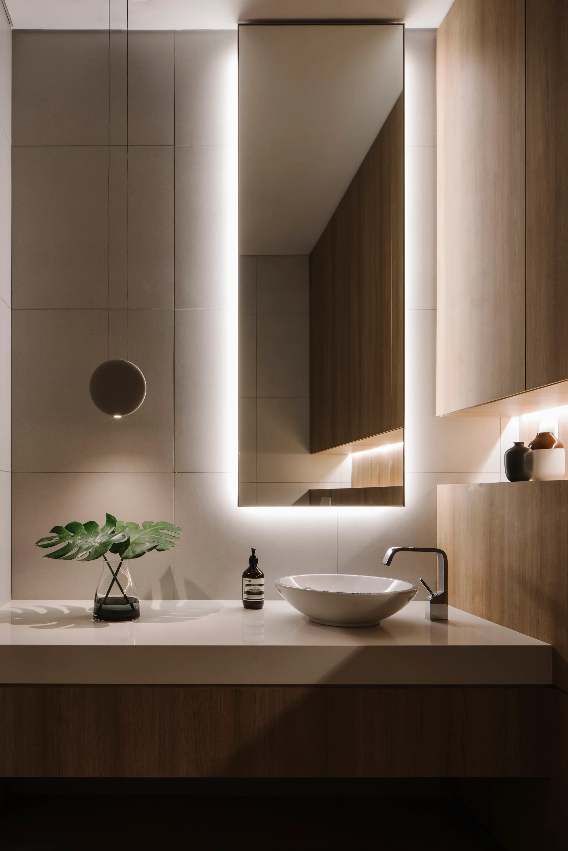 A minimalist bathroom vanity features a vessel sink, a tall backlit mirror, beige wall tiles, and wood cabinets.