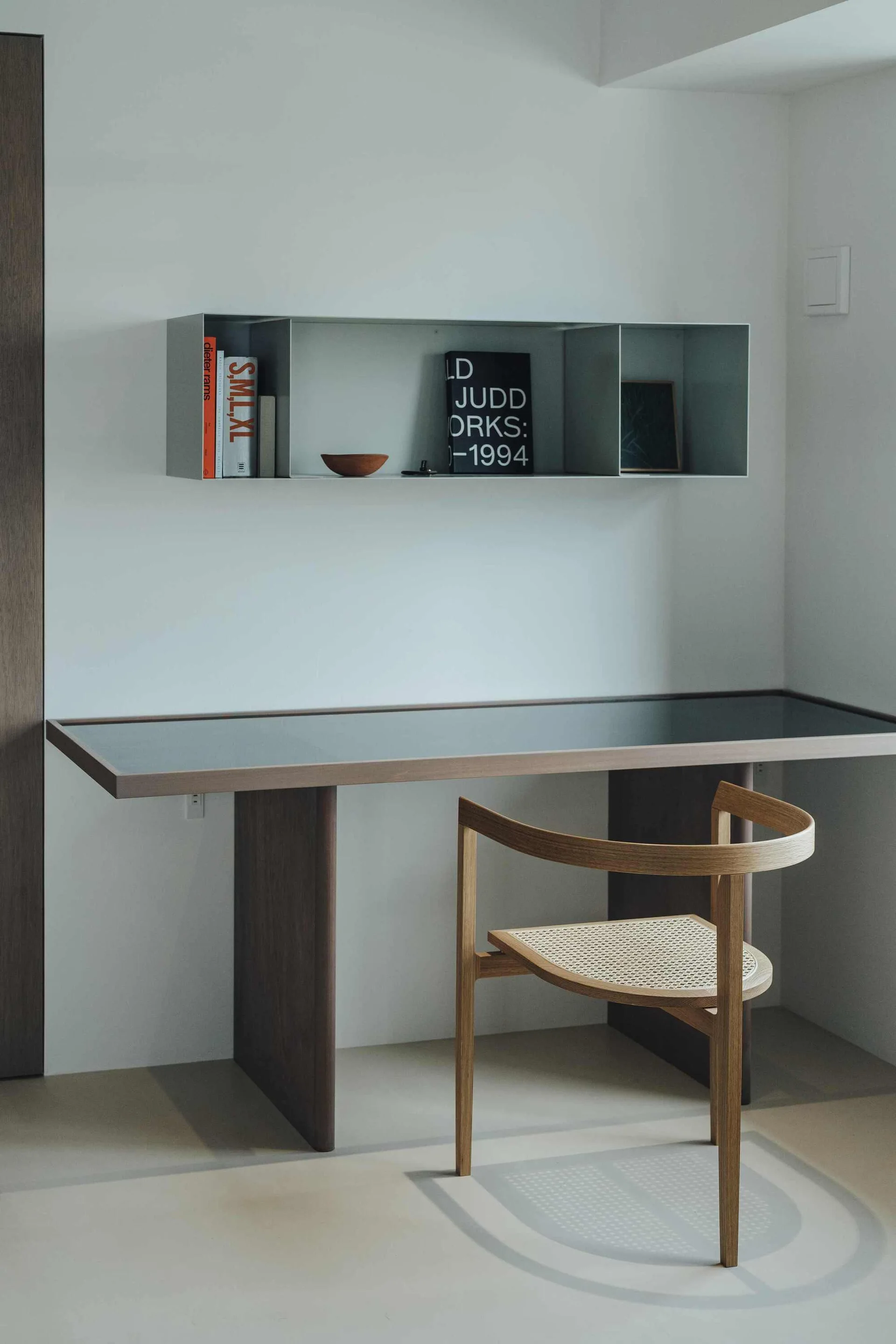A minimalist workspace features a dark wooden desk paired with a curved cane chair under a wall-mounted metal shelf holding books.