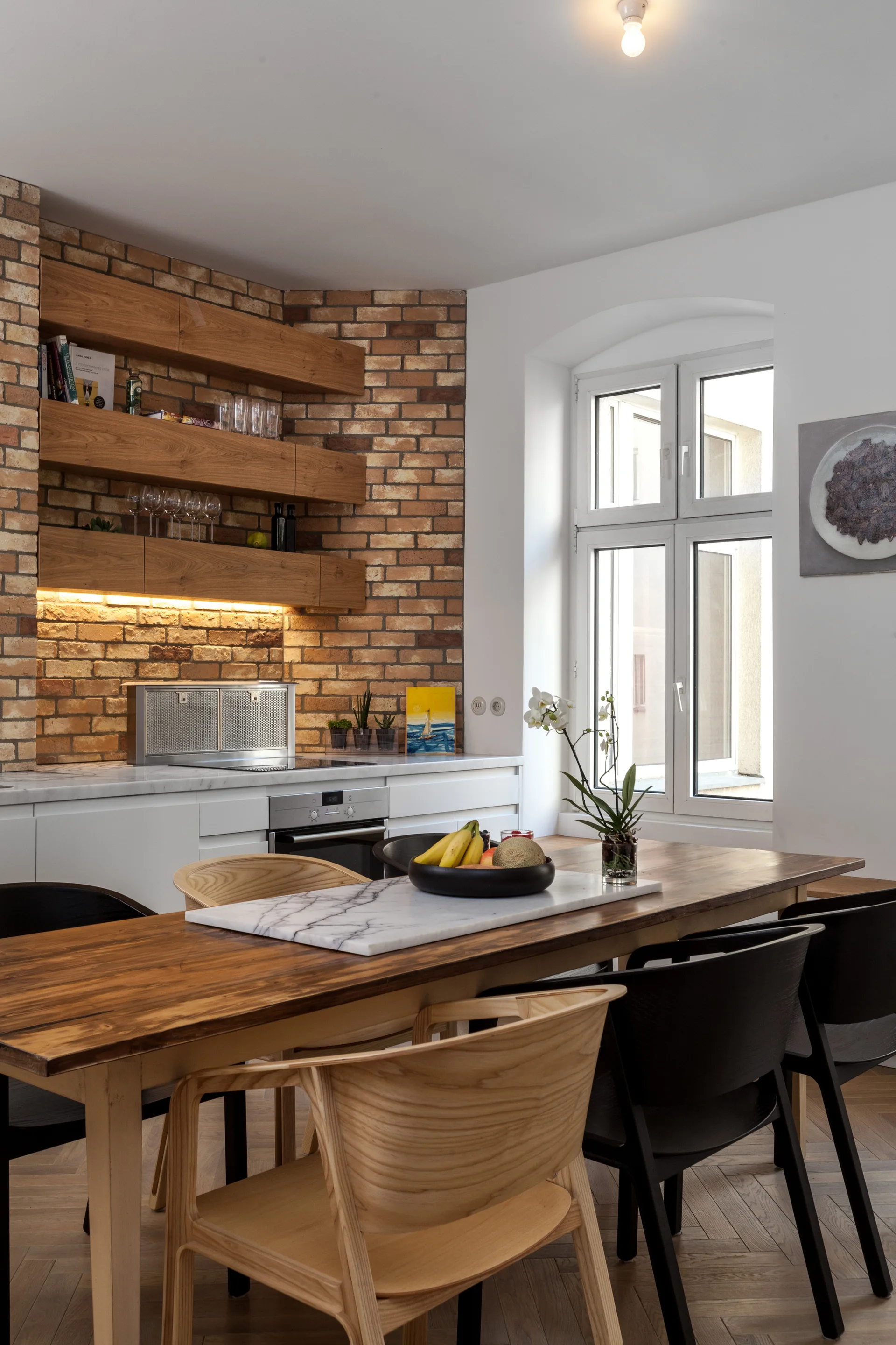 A wooden dining table sits in front of a white kitchen counter and an exposed brick accent wall with floating timber shelves.