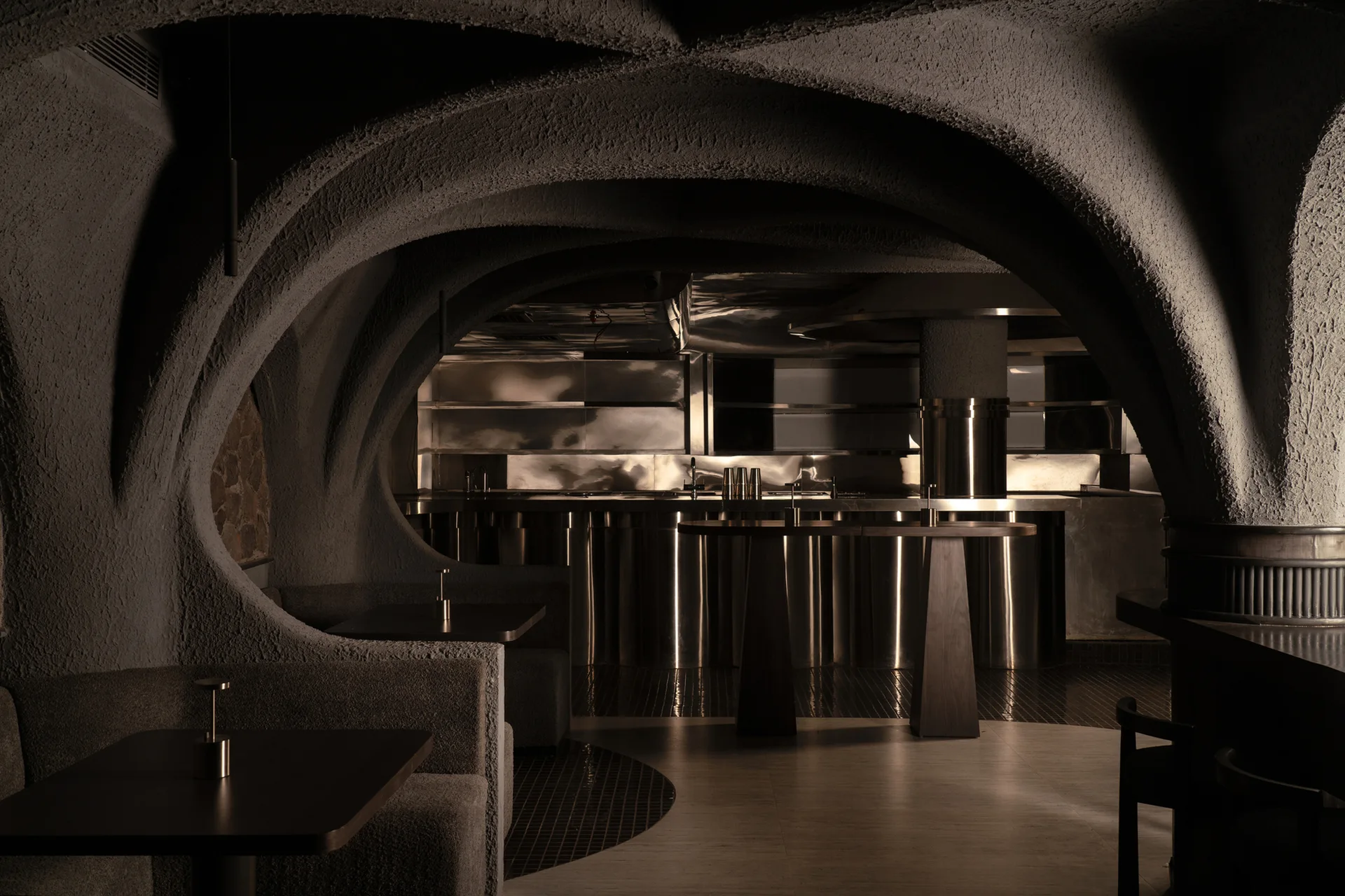 A dimly lit lounge features textured concrete vaulted arches, built-in upholstered booths, and a polished metallic bar counter in the background.