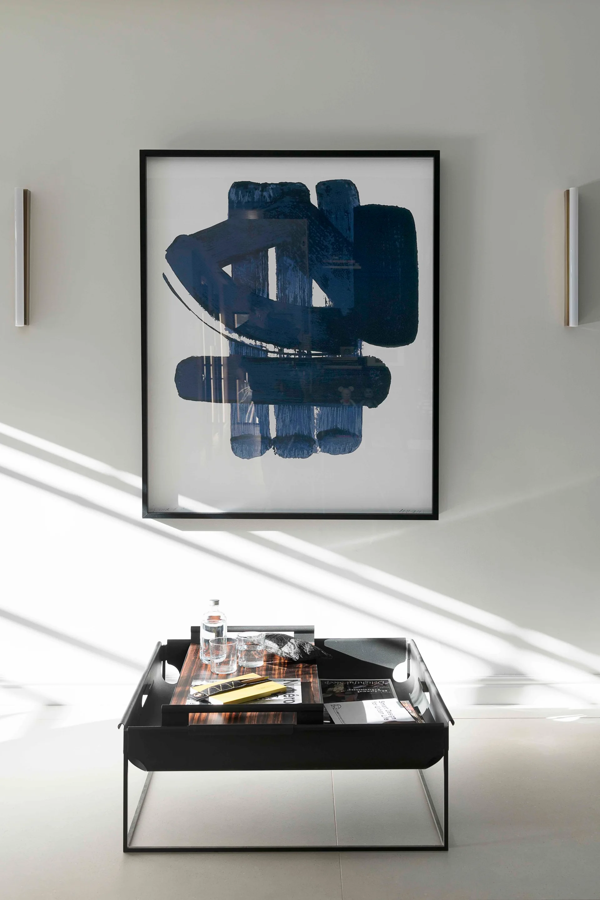 A framed blue abstract artwork hangs on a white wall between two slim vertical sconces above a black metal coffee table.