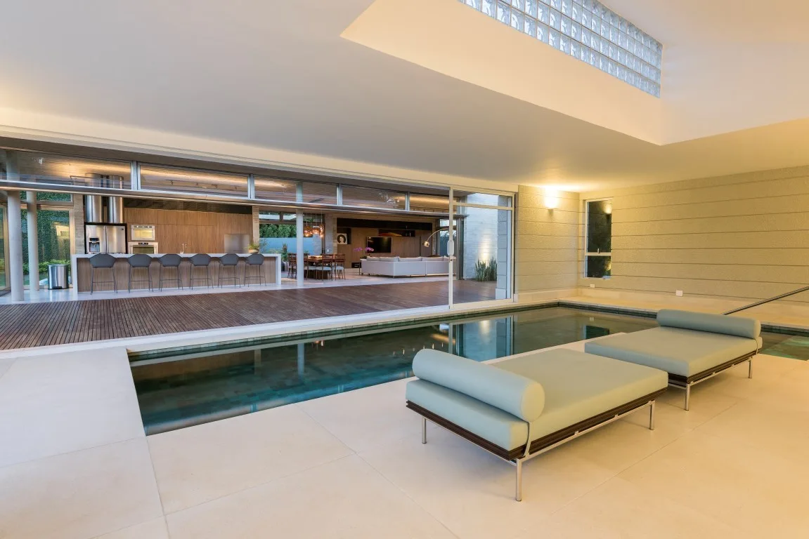 Two sage green daybeds sit beside an indoor swimming pool, which opens to a kitchen and living area through sliding glass doors.