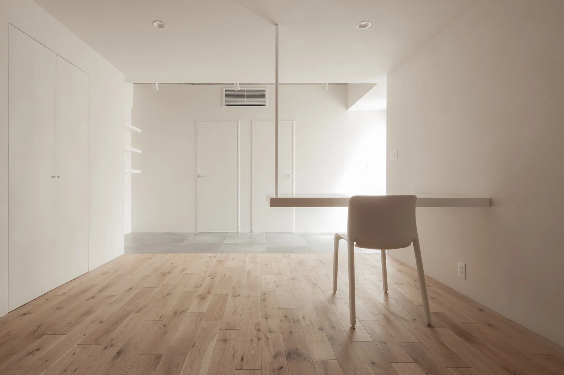 A minimalist room featuring a wall-mounted white table and chair on light wood flooring with white walls and doors in the background.