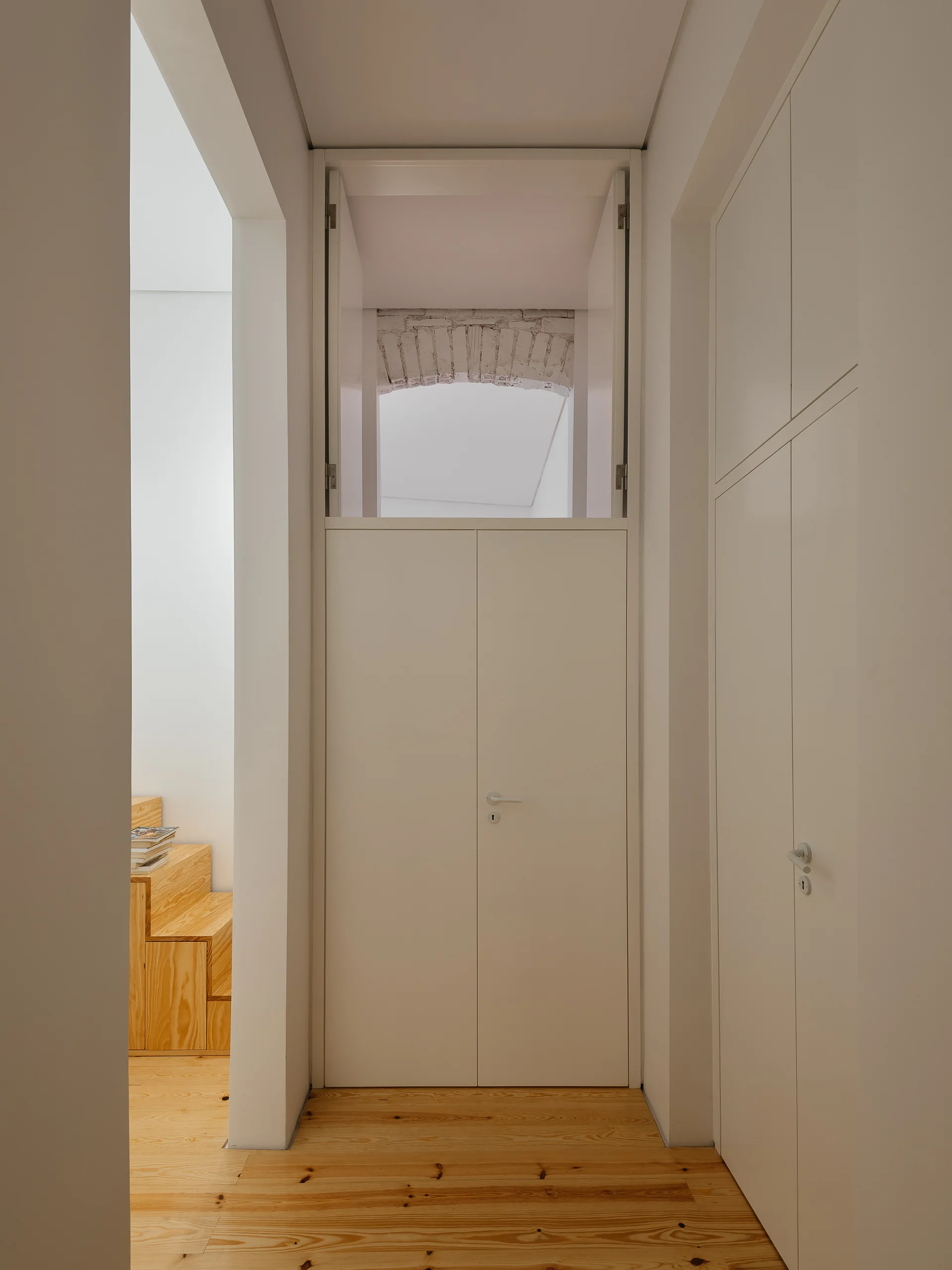 A minimalist white hallway features flush-fit built-in storage, light wood plank floors, and an exposed brick archway over a tall white door.