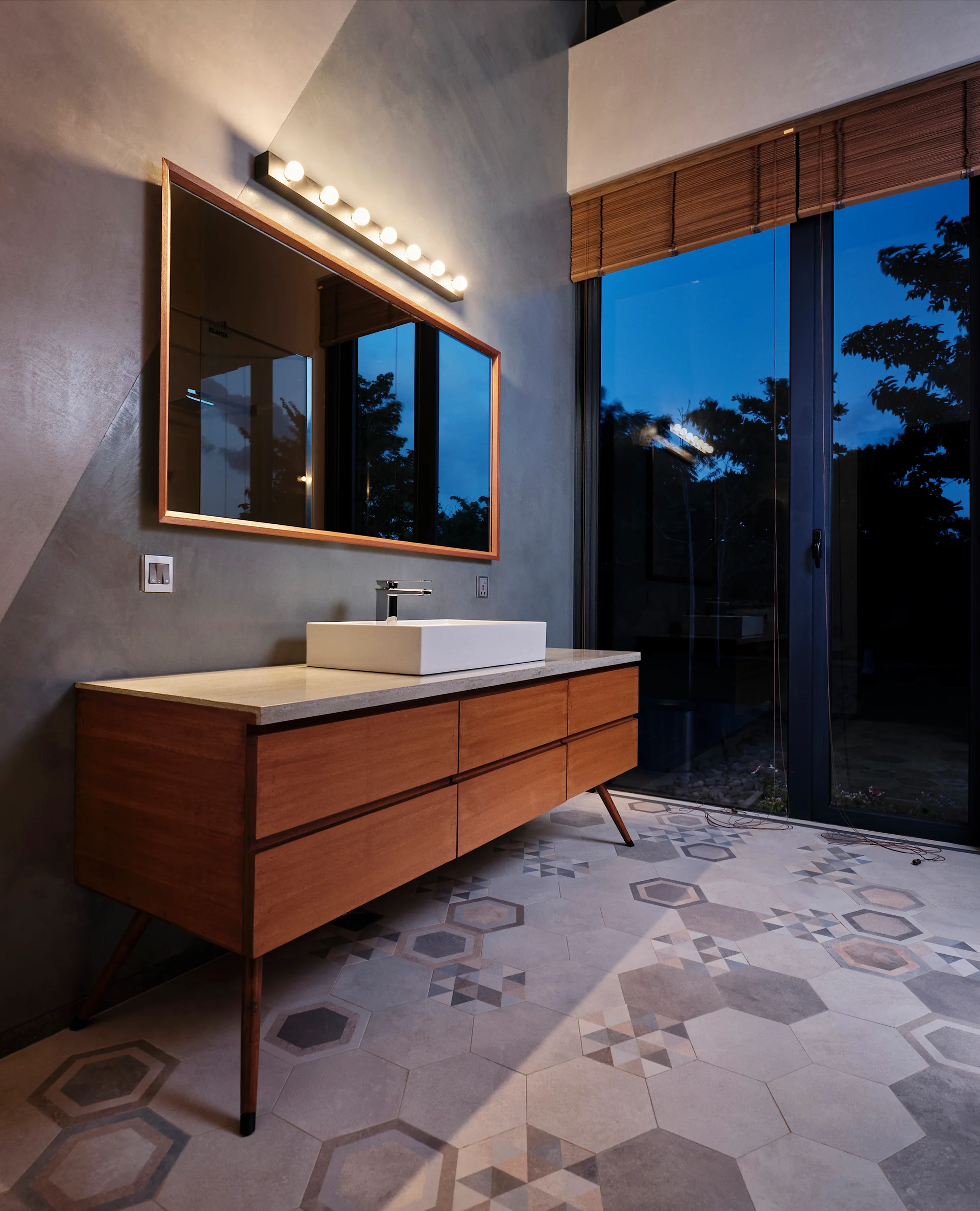 A modern bathroom features a warm wood vanity with a white vessel sink, a large wood-framed mirror, and grey hexagonal patterned floor tiles.