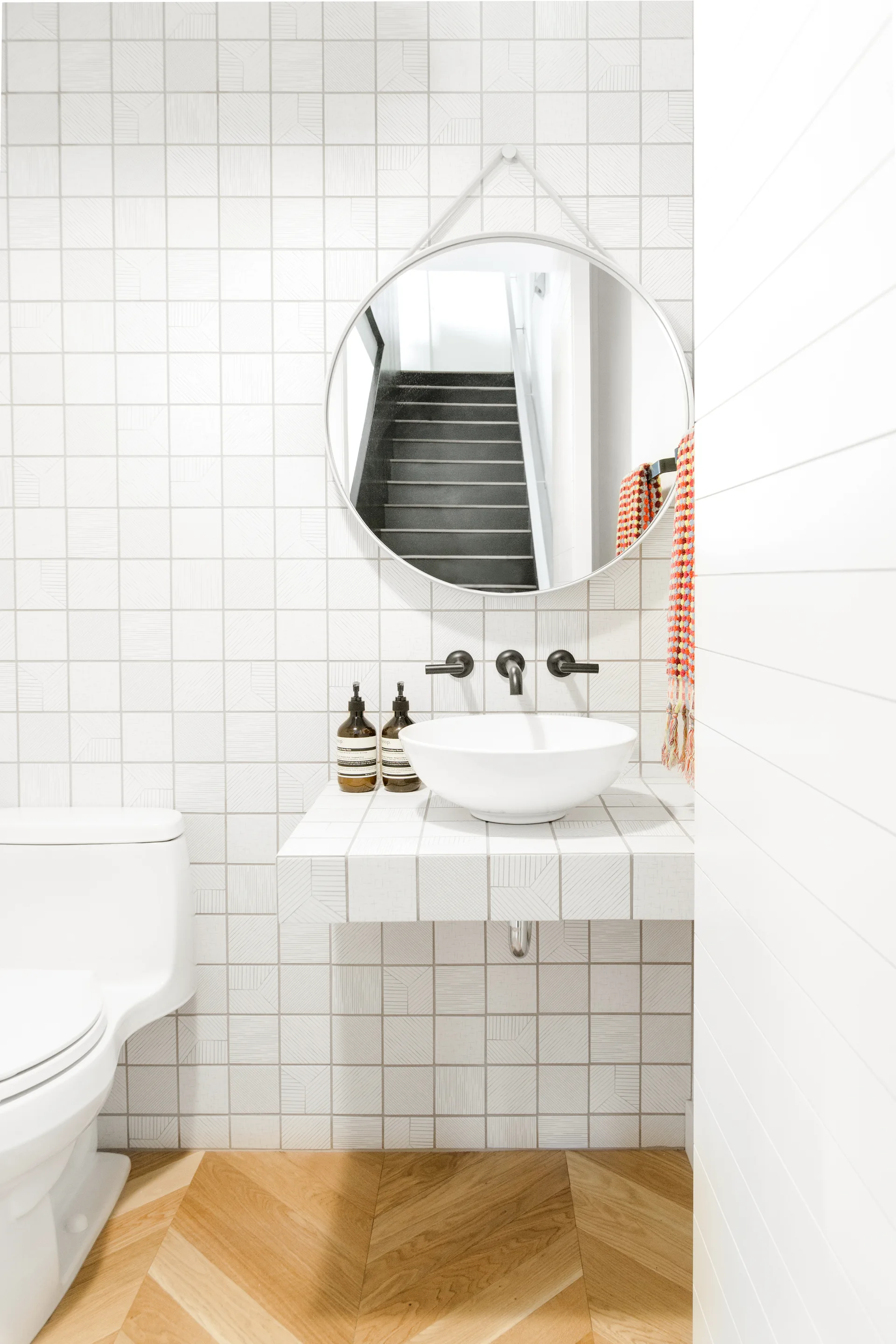 A modern powder room with white grid-tiled walls, a matching floating vanity, a round mirror, and a light wood herringbone floor.