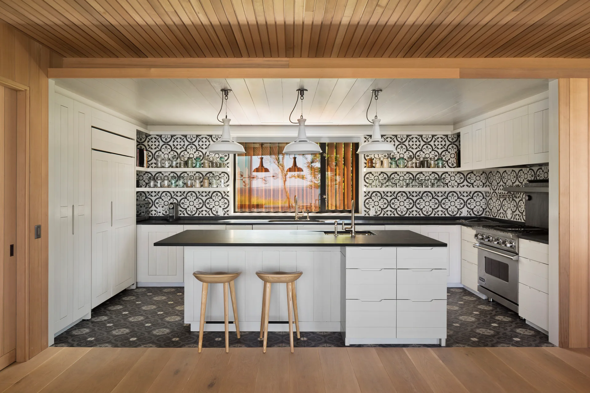 A modern kitchen featuring a white central island, two wooden bar stools, black countertops, a patterned tile backsplash, and a warm wood plank ceiling.