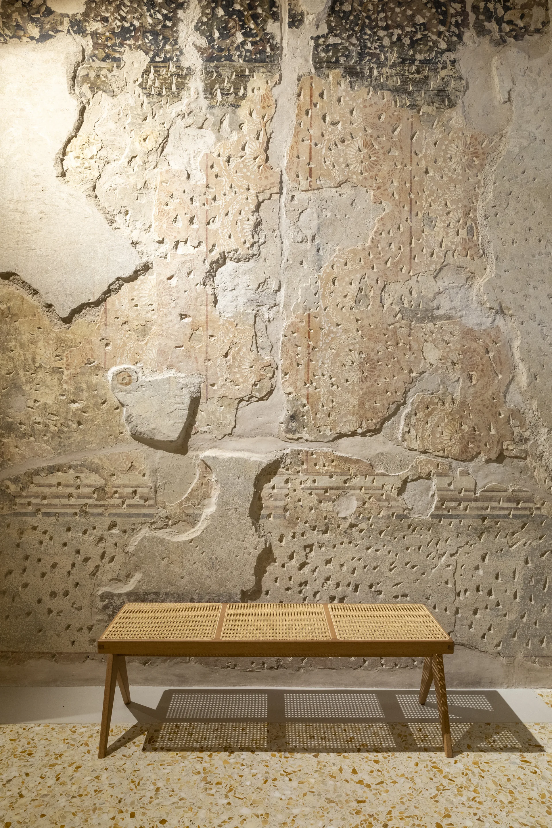 A wooden bench with a woven cane seat sits against a highly distressed beige plaster wall above a speckled terrazzo tile floor.