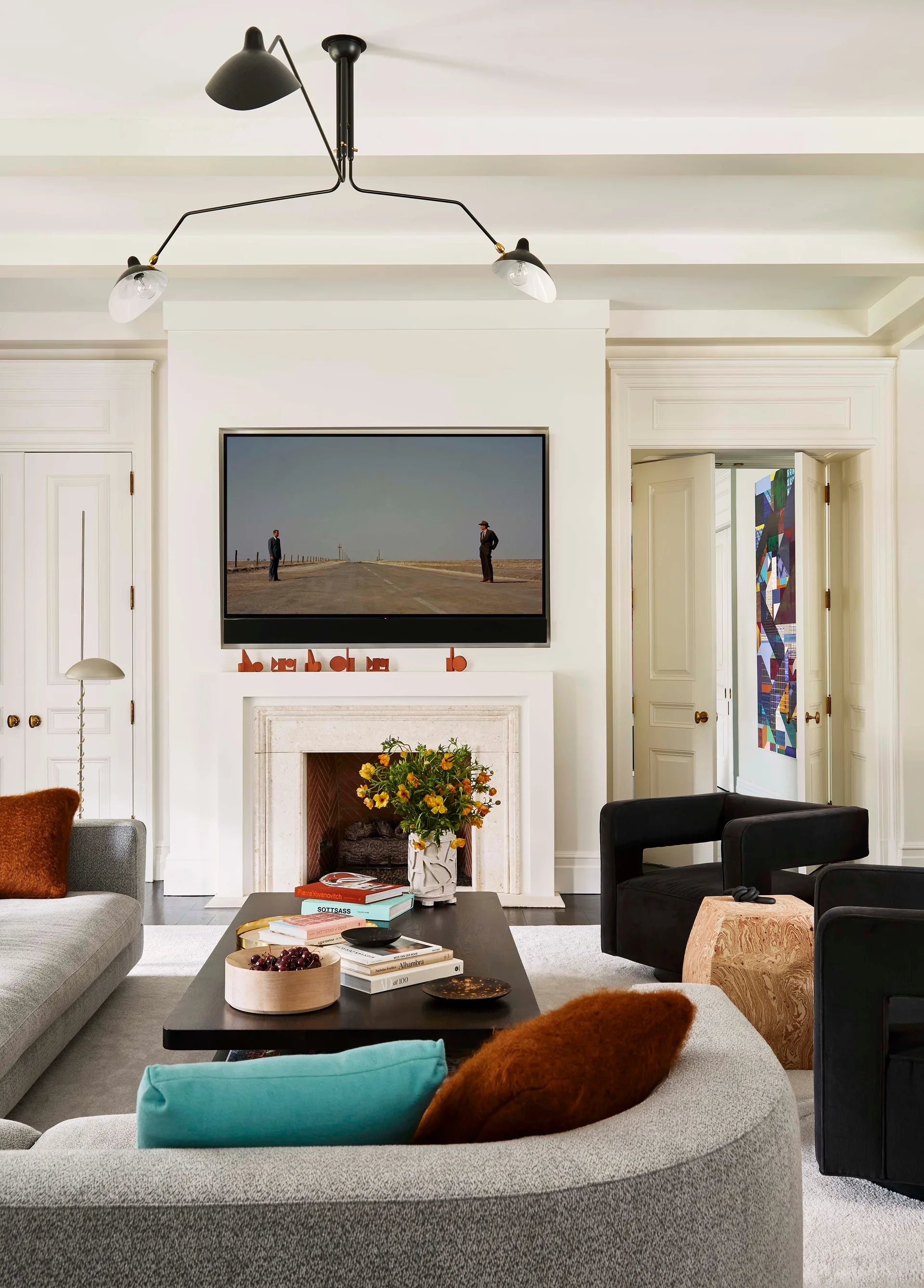 A living room features a stone fireplace with a TV above it, a curved gray sofa, two black armchairs, and a black multi-arm ceiling light.