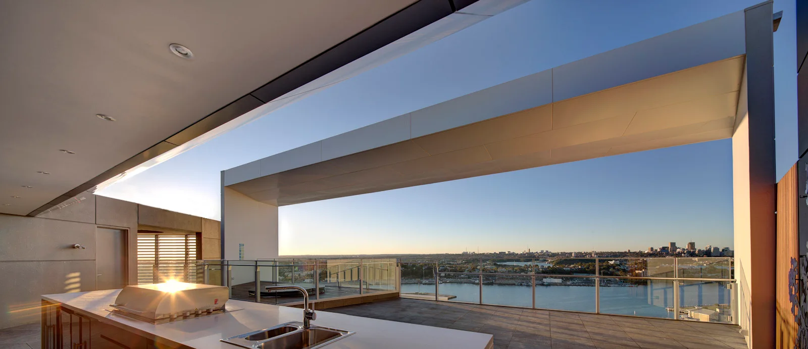 A modern outdoor kitchen counter with a stainless steel grill and sink sits on a spacious tiled terrace with glass railings overlooking a city waterfront.